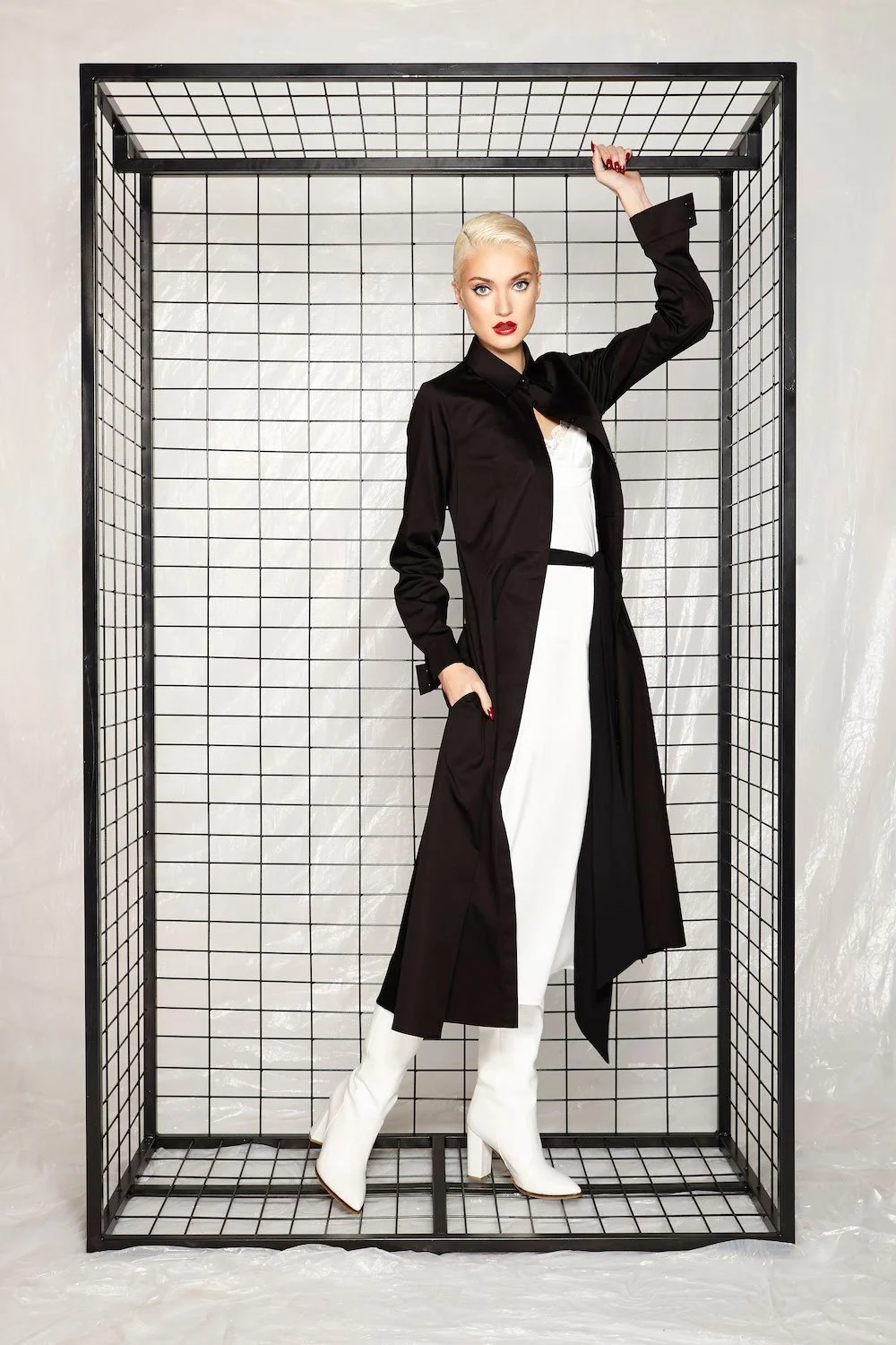 A fashionable woman with platinum blonde hair, red lipstick, and striking makeup poses inside a black wire frame structure. She is wearing a long black coat, white pants, and white knee-high boots with heels.
