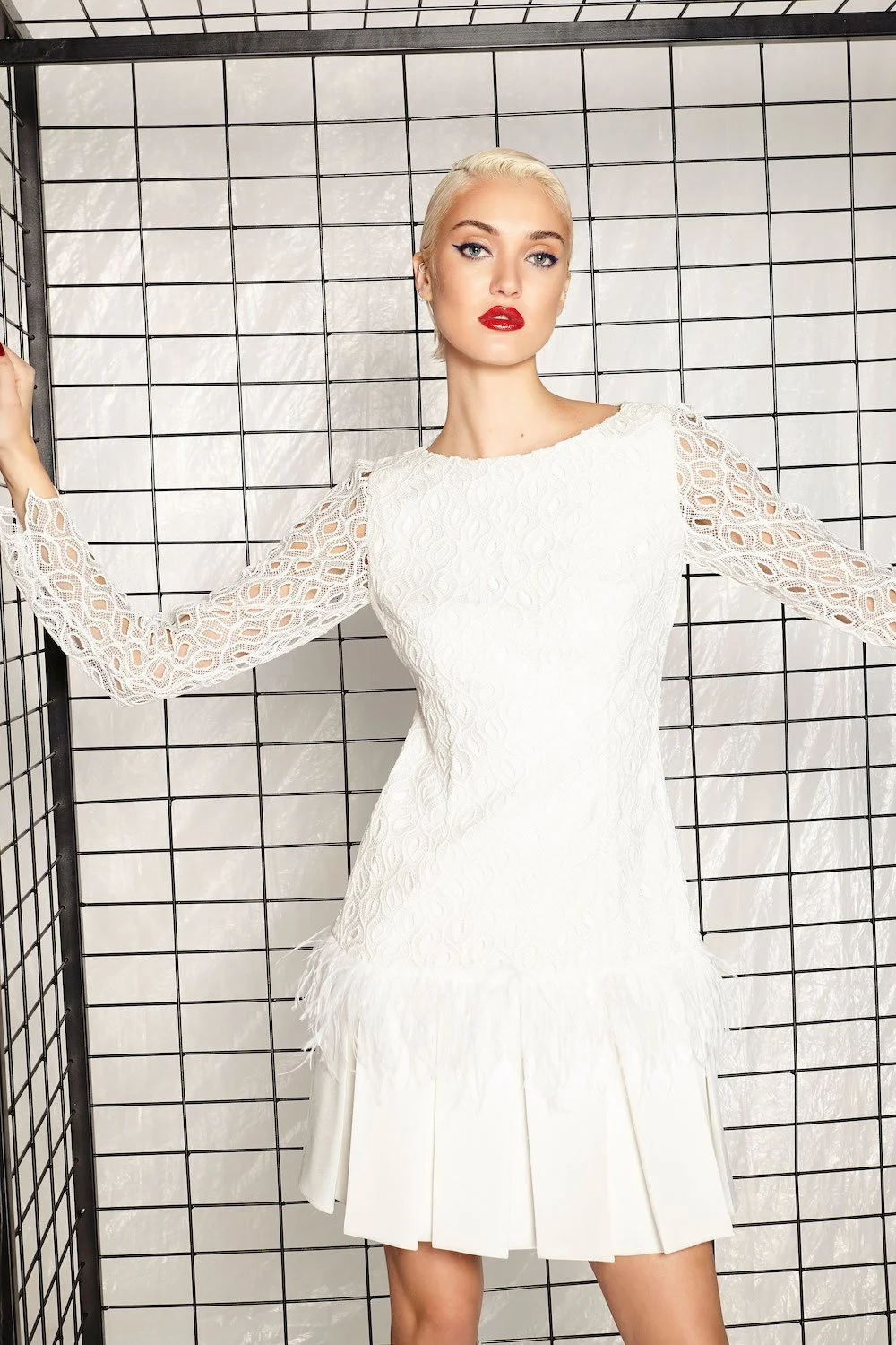 A woman with platinum blonde hair and red lipstick wearing a white dress with ovoid-patterned, open-knit sleeves and a feathered, pleated skirt standing against a black wire grid backdrop.