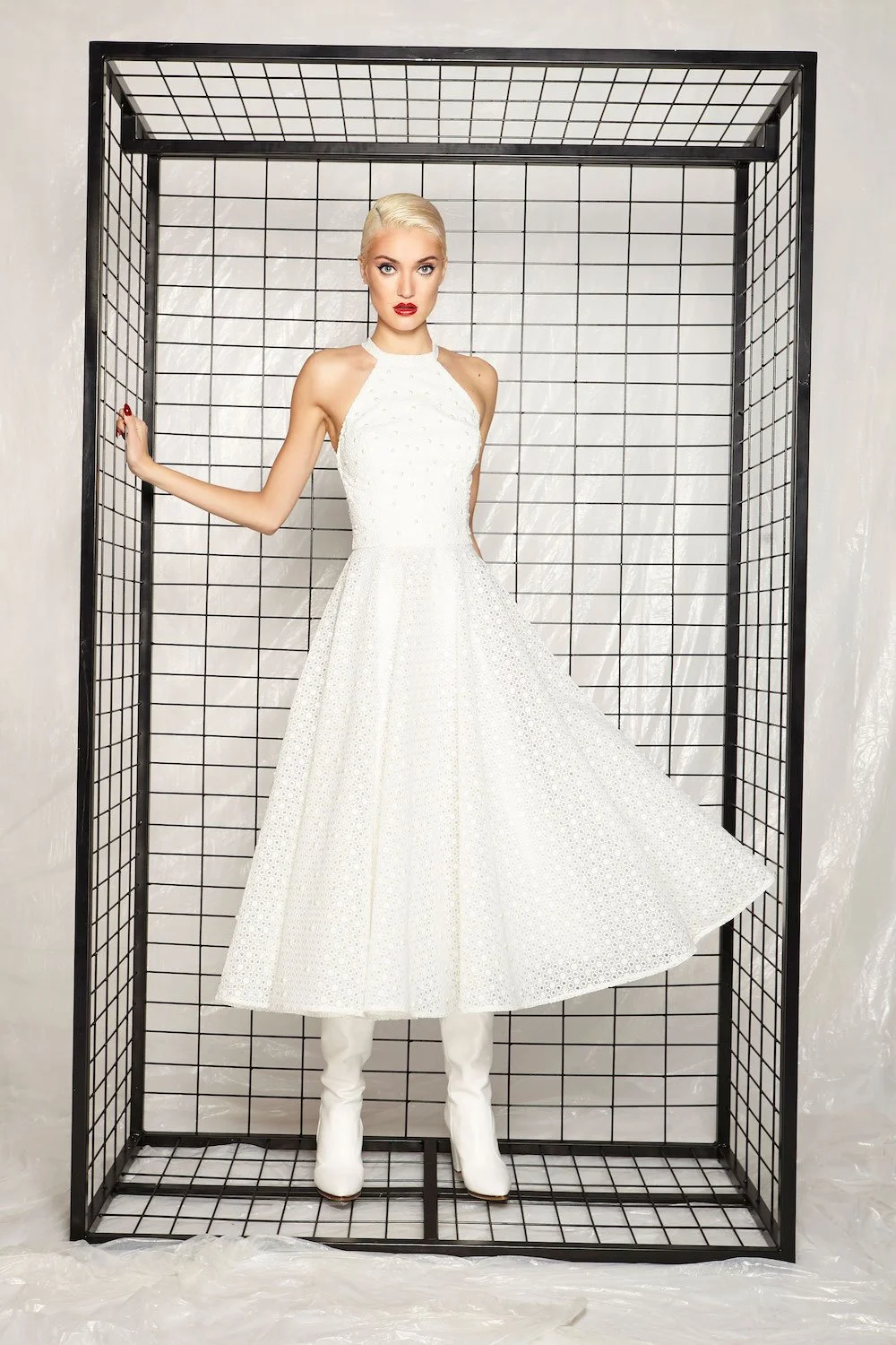 A woman with platinum blonde hair and red lipstick posing inside a metal wireframe enclosure, wearing a white sleeveless dress and matching white boots.