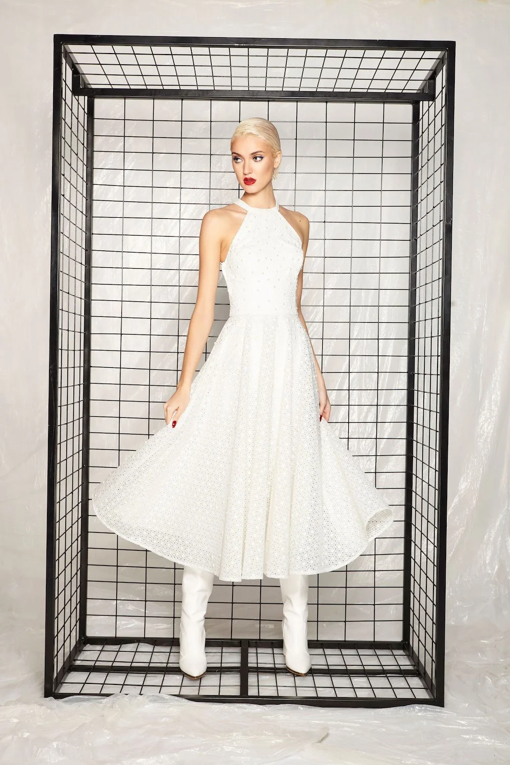 A woman with platinum blonde hair in a white dress and white boots standing inside a black metal grid structure.