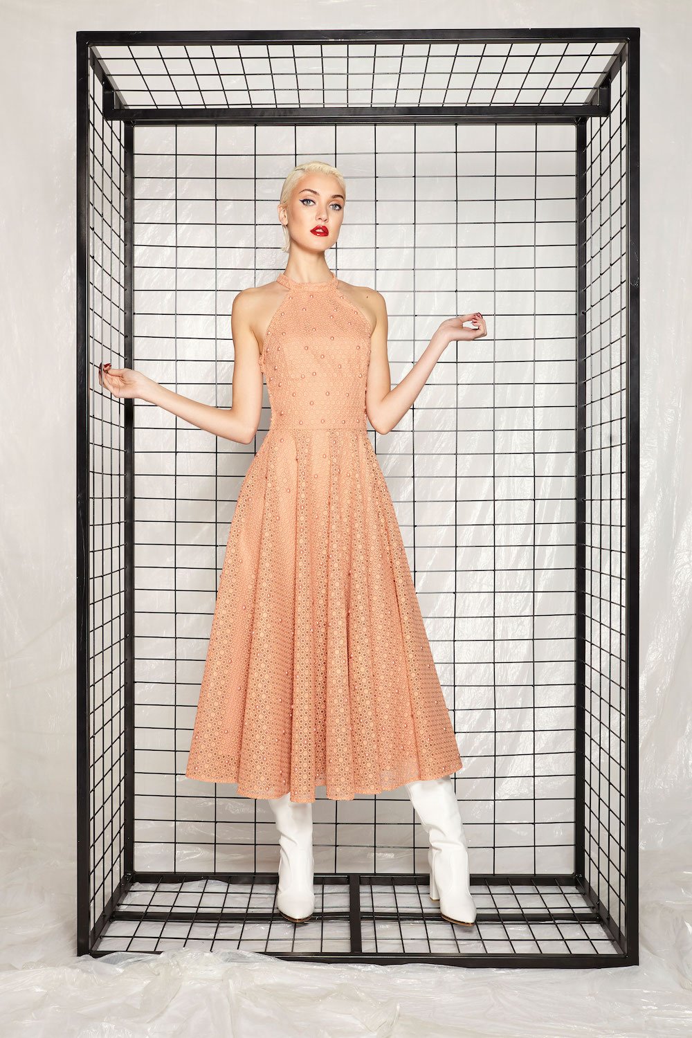 Woman in peach-colored dress and white boots standing inside a black metal grid enclosure.