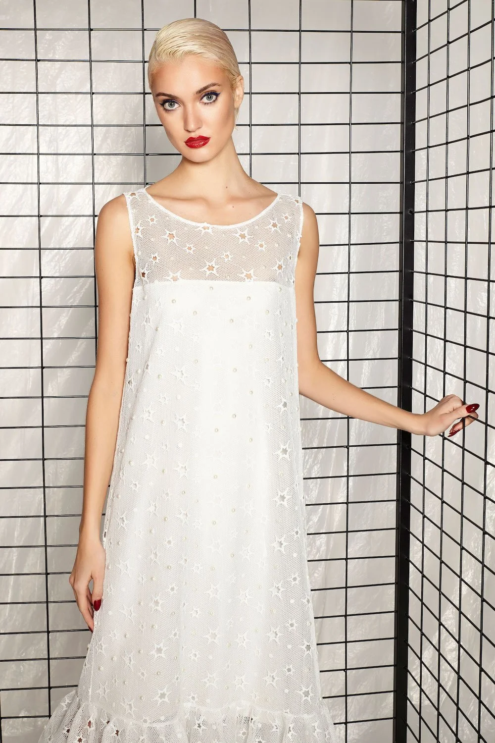 A young woman with platinum blonde hair, red lipstick, and blue eyes wearing a sleeveless white dress with star-patterned mesh, standing against a black wire grid backdrop.