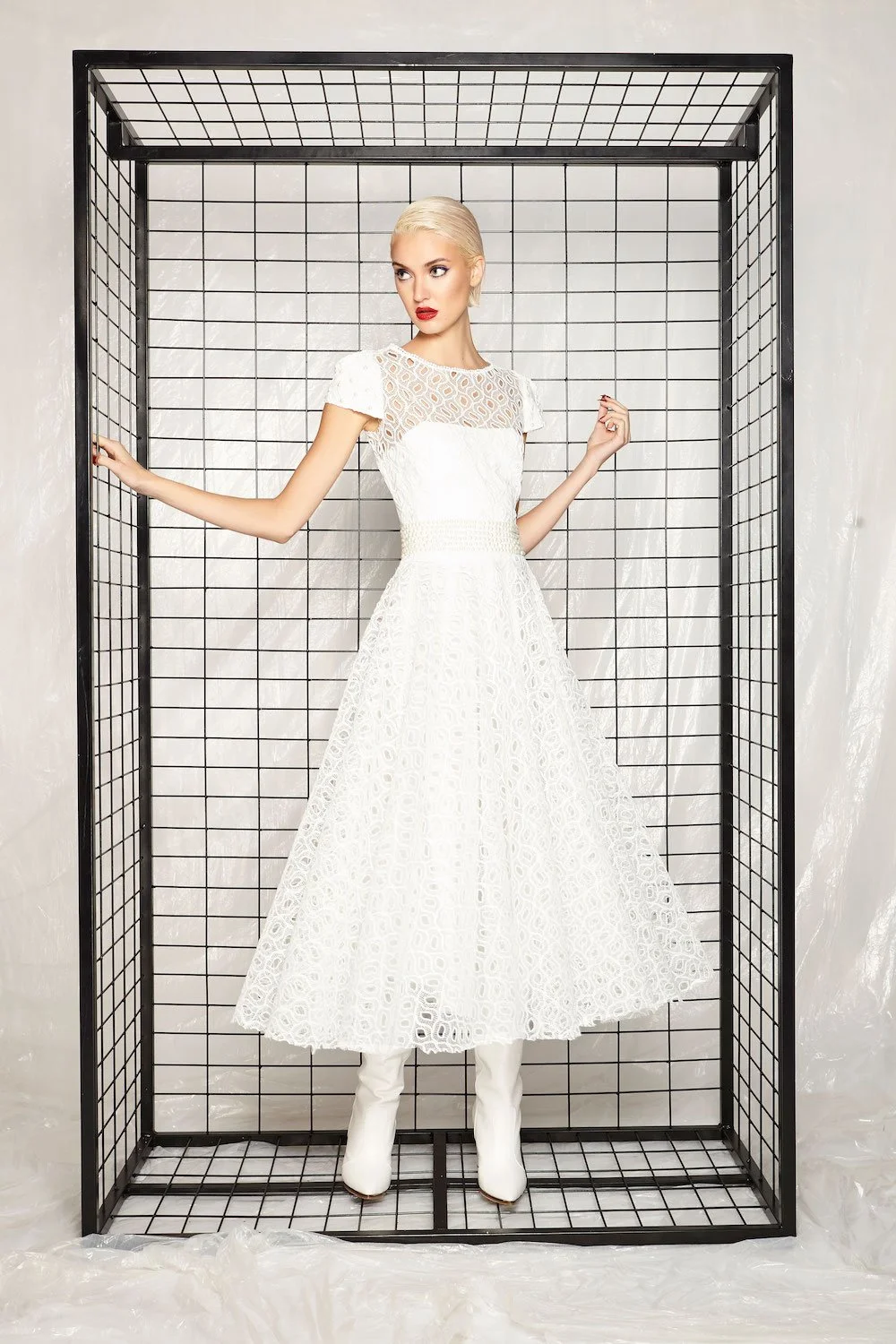 A woman with platinum blonde hair, red lipstick, and glamorous makeup posing in a white lace dress with a high waist and puffed sleeves, standing inside a black wire cube against a plain background.