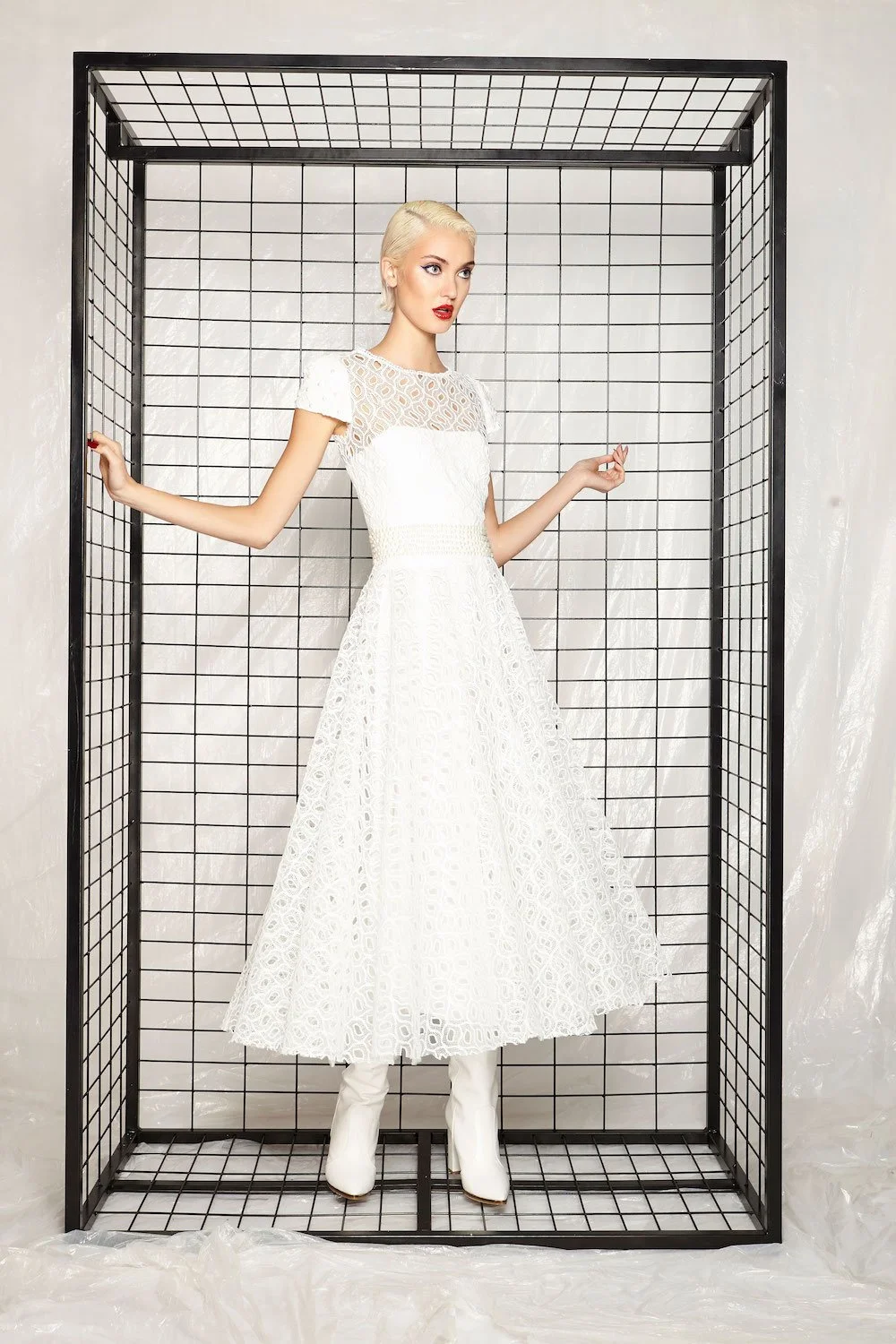 A woman with platinum blonde hair wearing a white dress and white boots posing inside a black grid structure.
