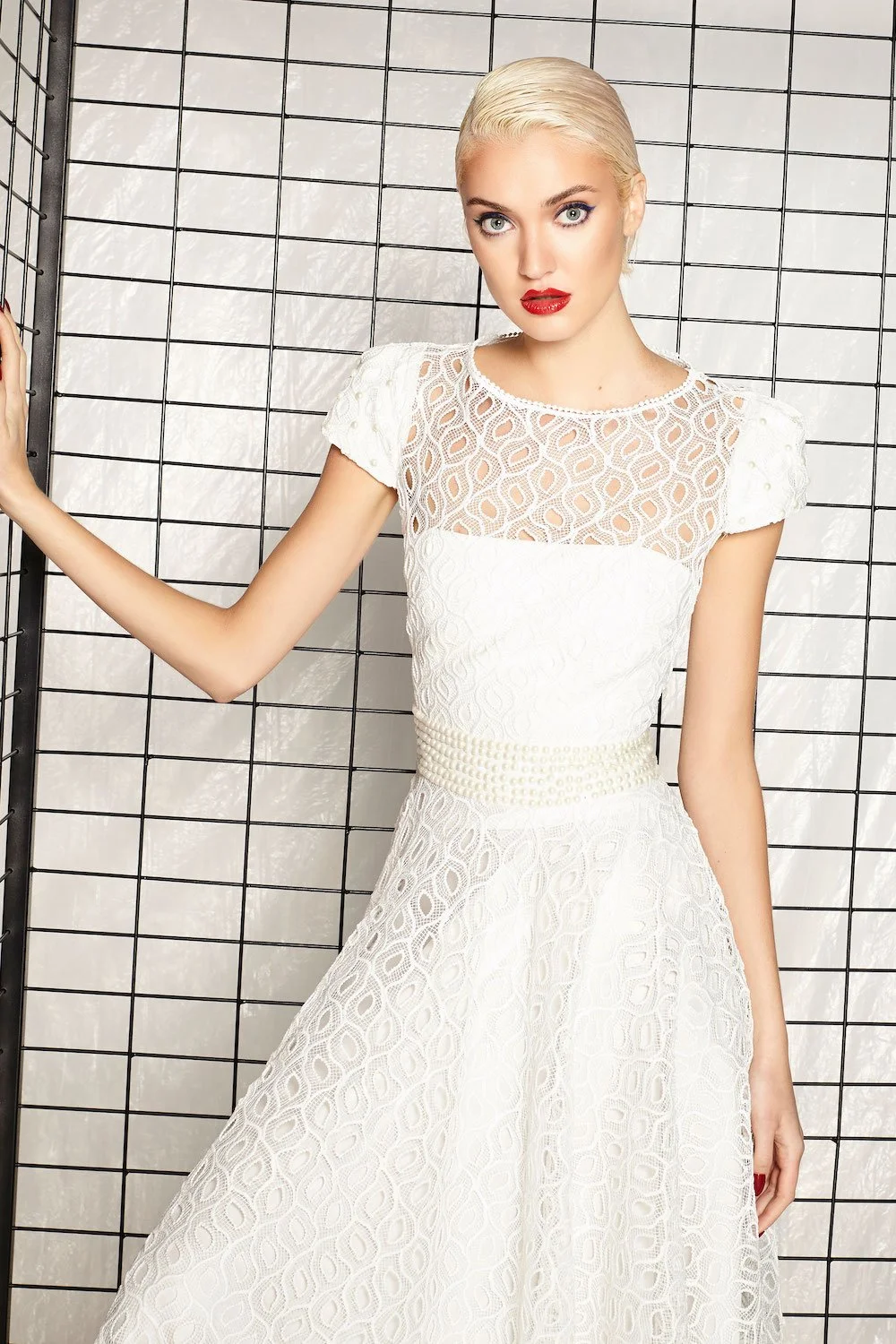 A woman with platinum blonde hair styled back, wearing bold makeup with red lipstick and blue eyeshadow, dressed in a white lace dress with a fitted waist and short sleeves, standing against a black wire grid background.