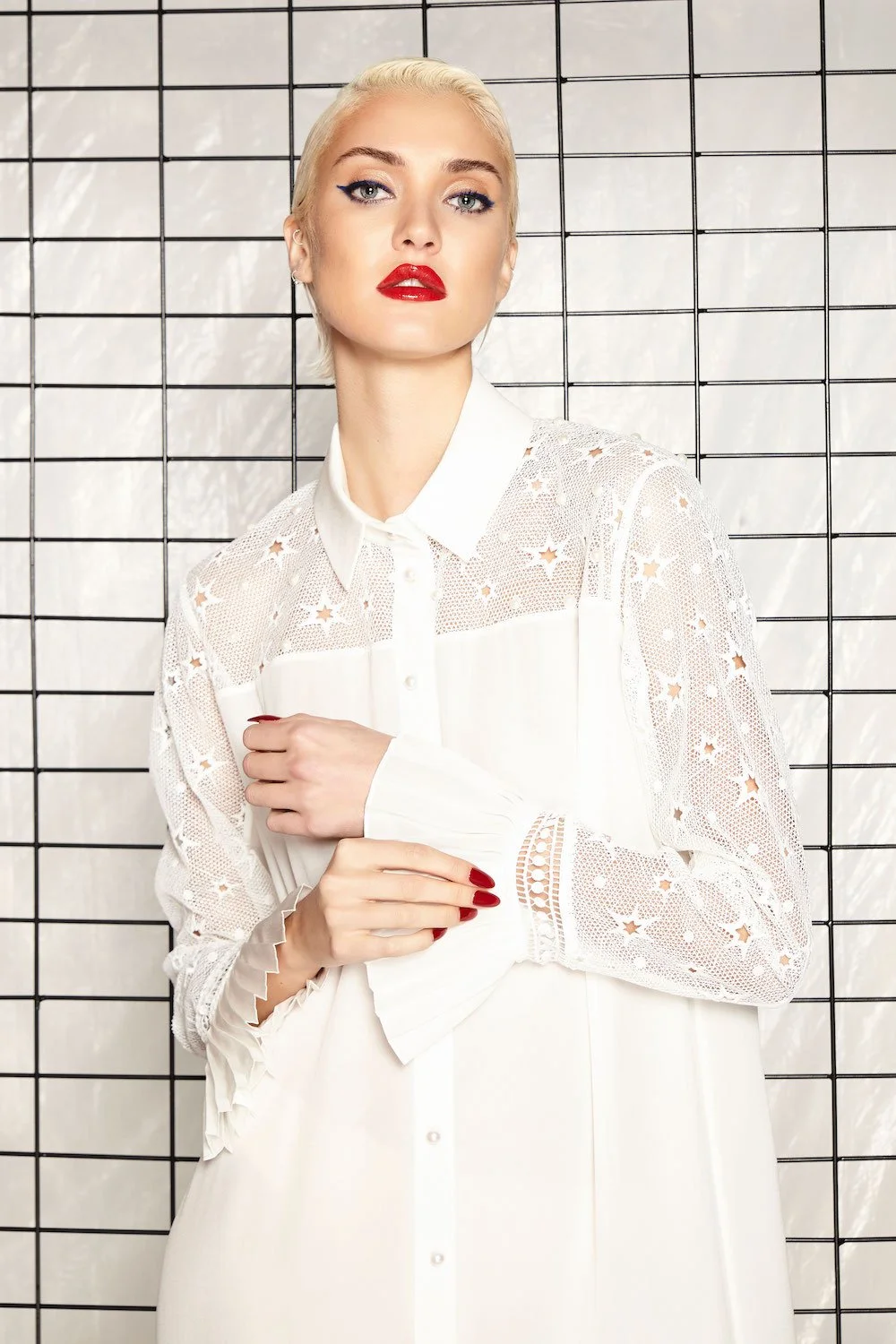 A woman with platinum blonde hair and red lipstick posing in front of a white tiled wall. She is wearing a white, long-sleeved dress with lace and star-shaped cutouts, and has manicured nails painted red.