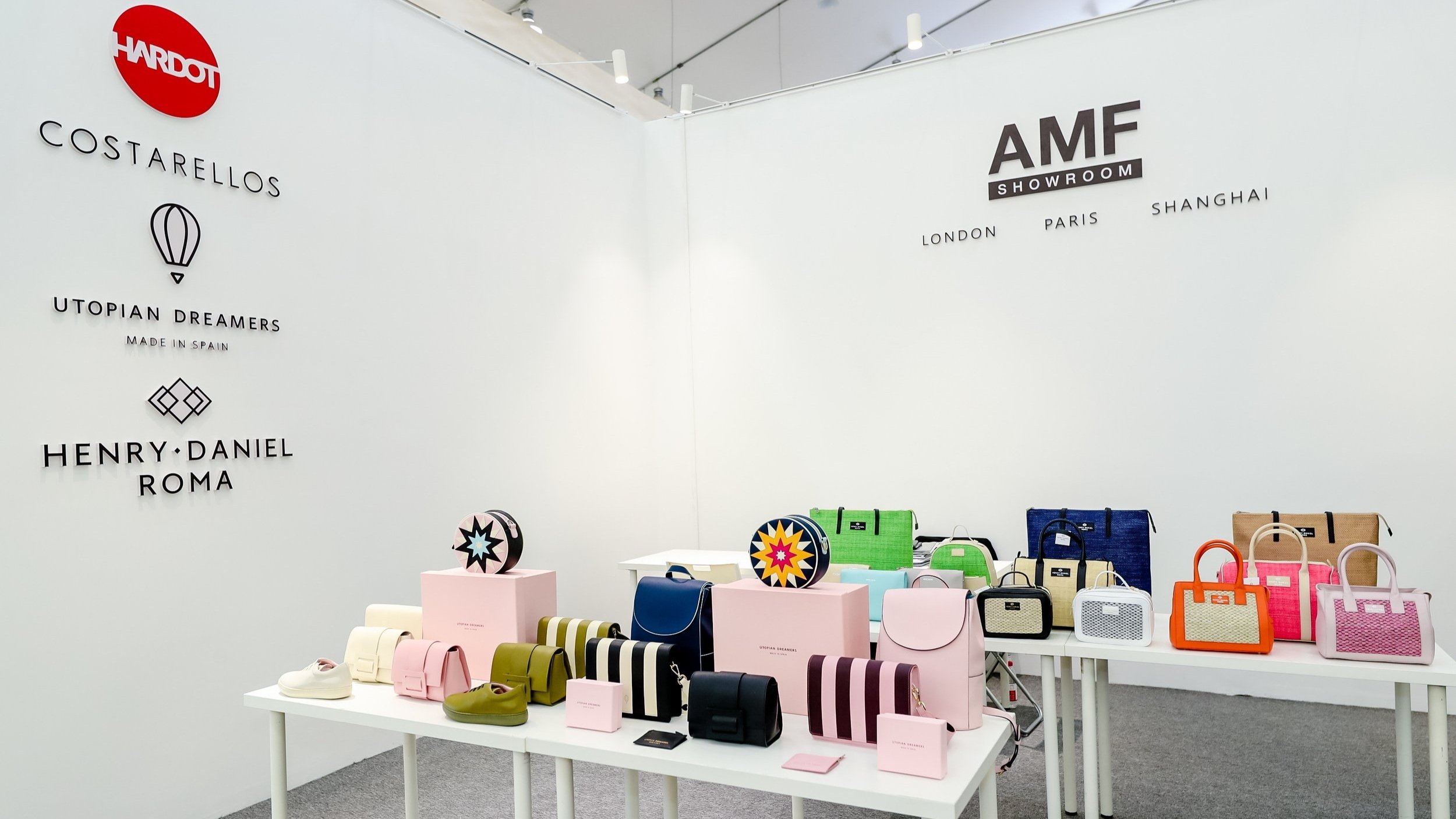Display of colorful handbags, shoes, and accessories on white tables at a showroom with logos and brand names on the white walls.