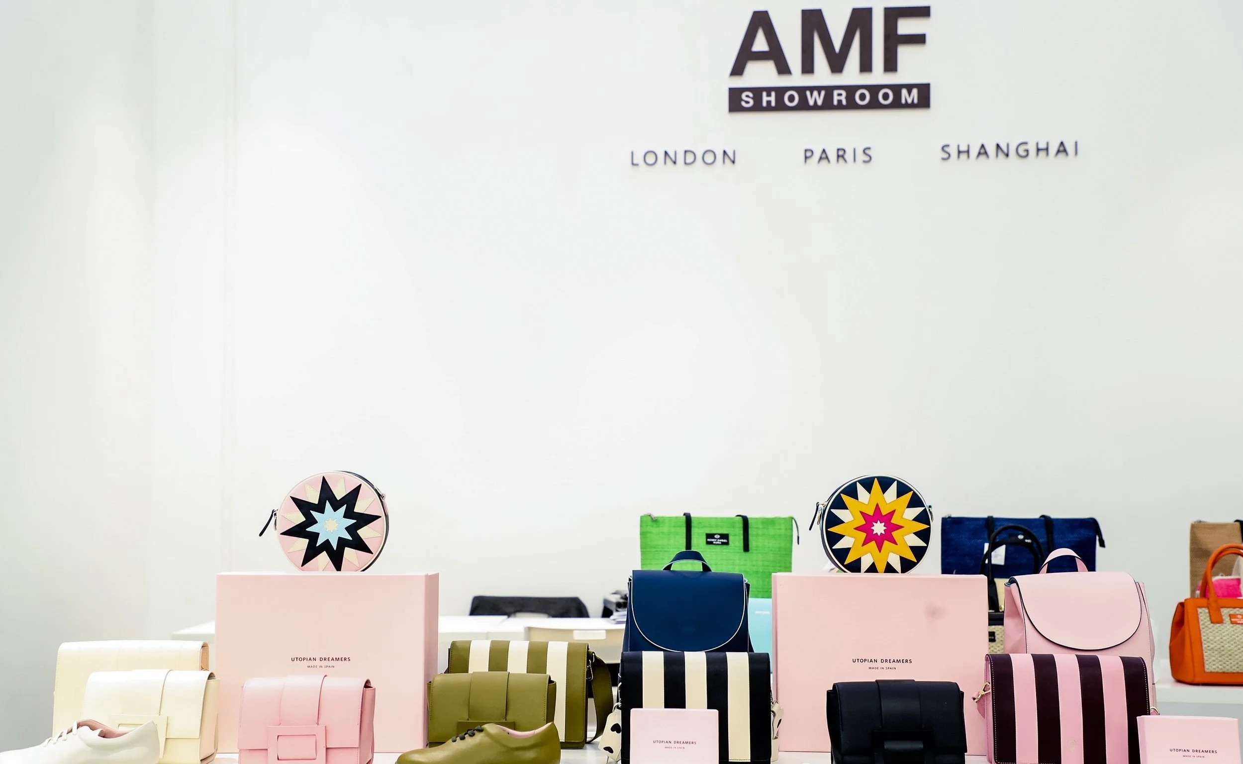 Display table with various handbags and shoes at a fashion showroom. The backdrop has the logo 'AMF SHOWROOM' and locations listed as London, Paris, and Shanghai.