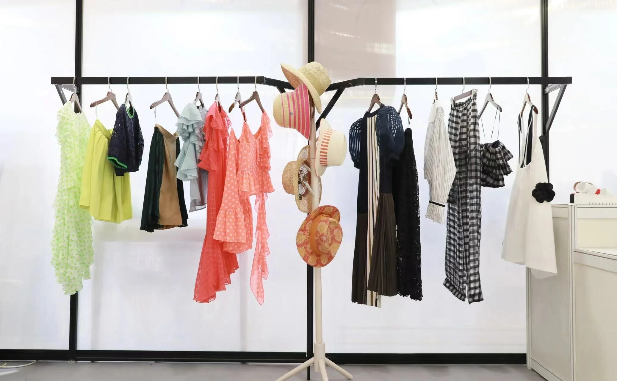 Clothing rack with colorful dresses, shirts, and skirts, and a stand with various hats. The background features large windows.