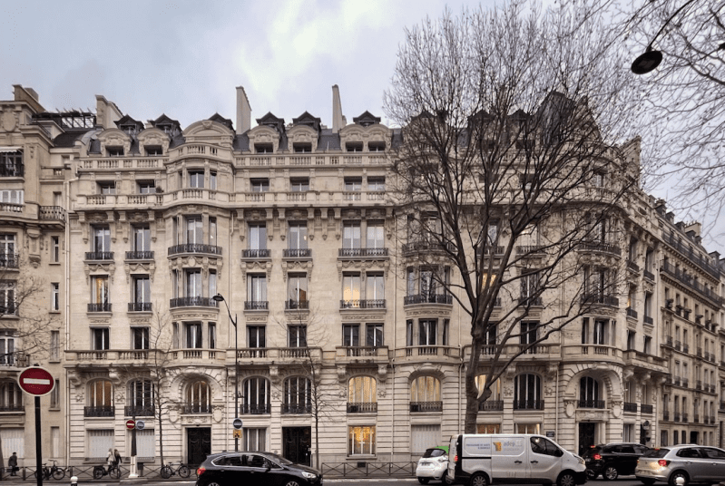 Street view of a classic European building with multiple floors, ornate details, and a tree in front. Several cars and bicycles are parked along the street, with a 'do not enter' sign visible.
