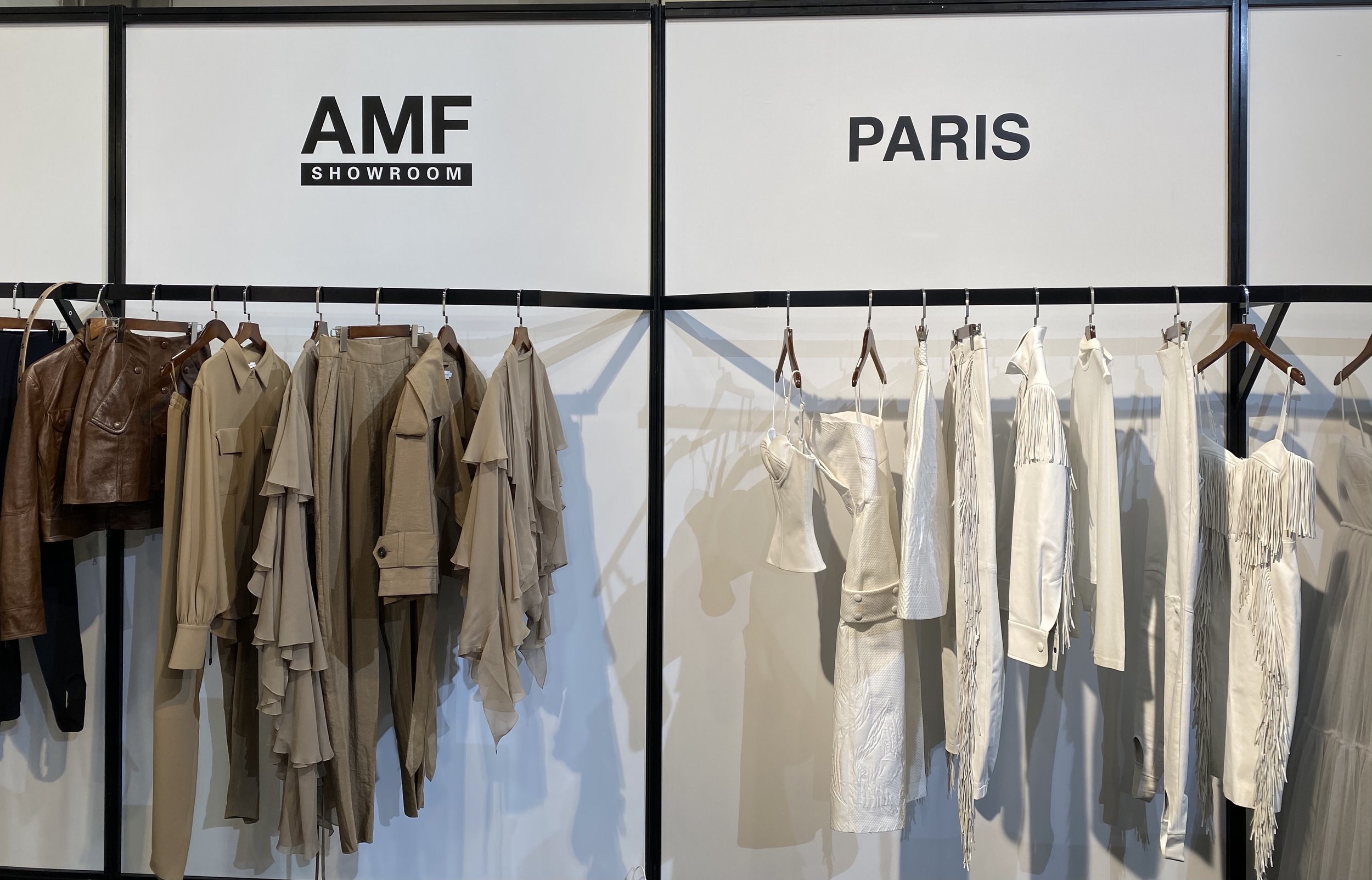 Clothing display with beige and white garments on racks, labeled 'AMF SHOWROOM PARIS' in black letters.