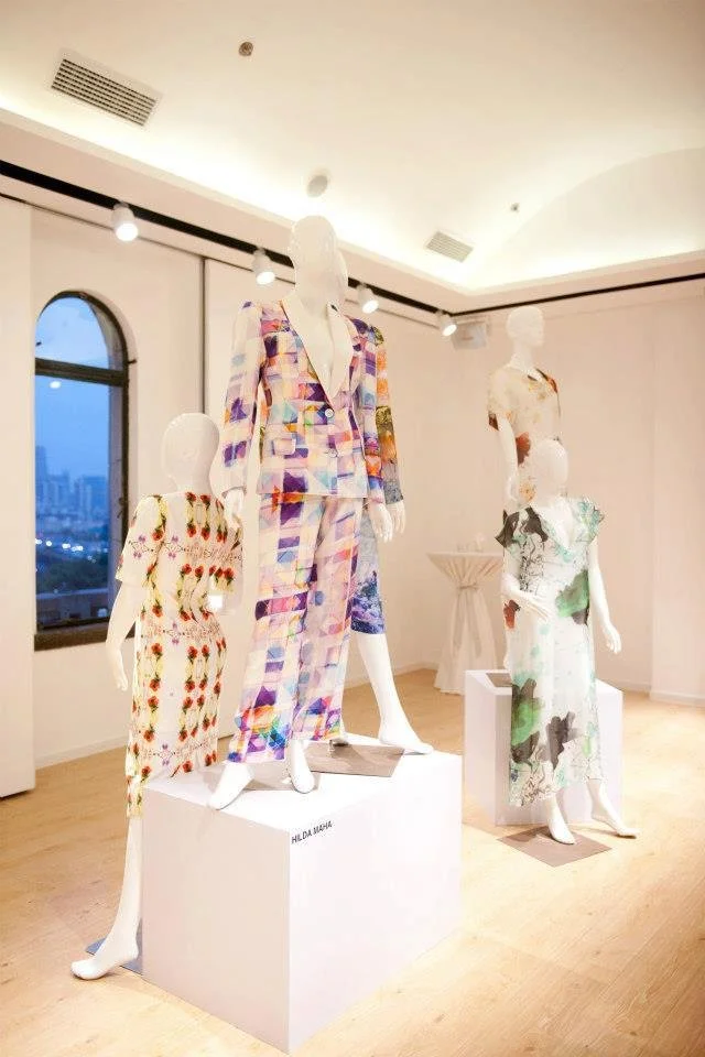 Display of four mannequins dressed in colorful, patterned designer clothing showcased on white pedestals in an art gallery or showroom with a city view through the window.