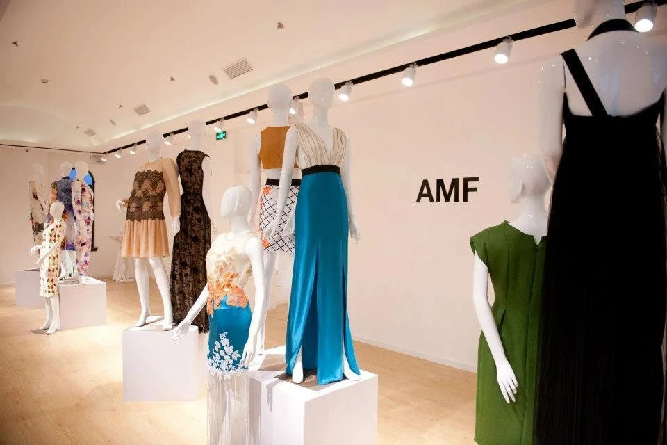 Display of mannequins wearing colorful dresses in an art gallery or fashion exhibit with the letters 'AMF' on the wall.