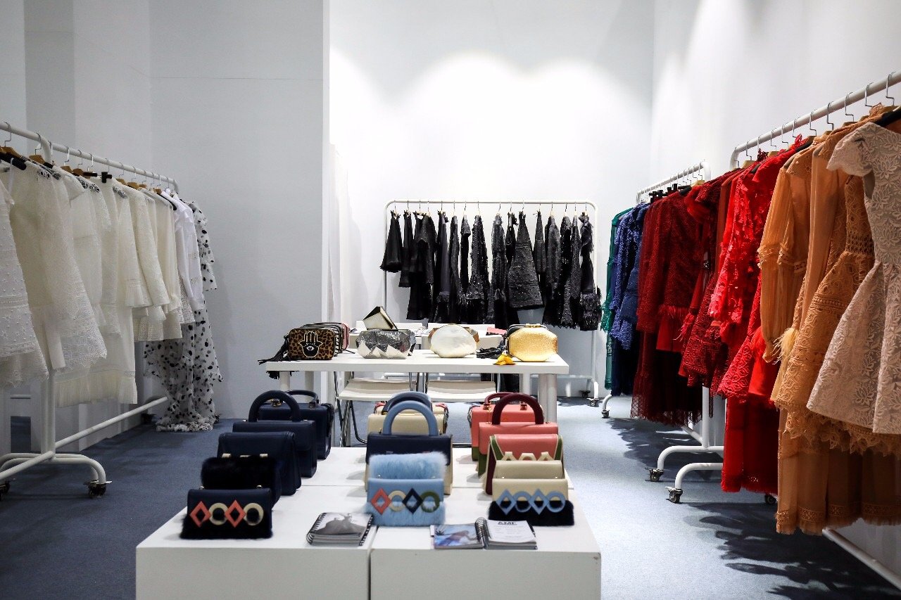 Fashion boutique with clothing racks, handbags, and accessories displayed on tables and racks, featuring dresses in various colors including white, black, red, green, and beige.