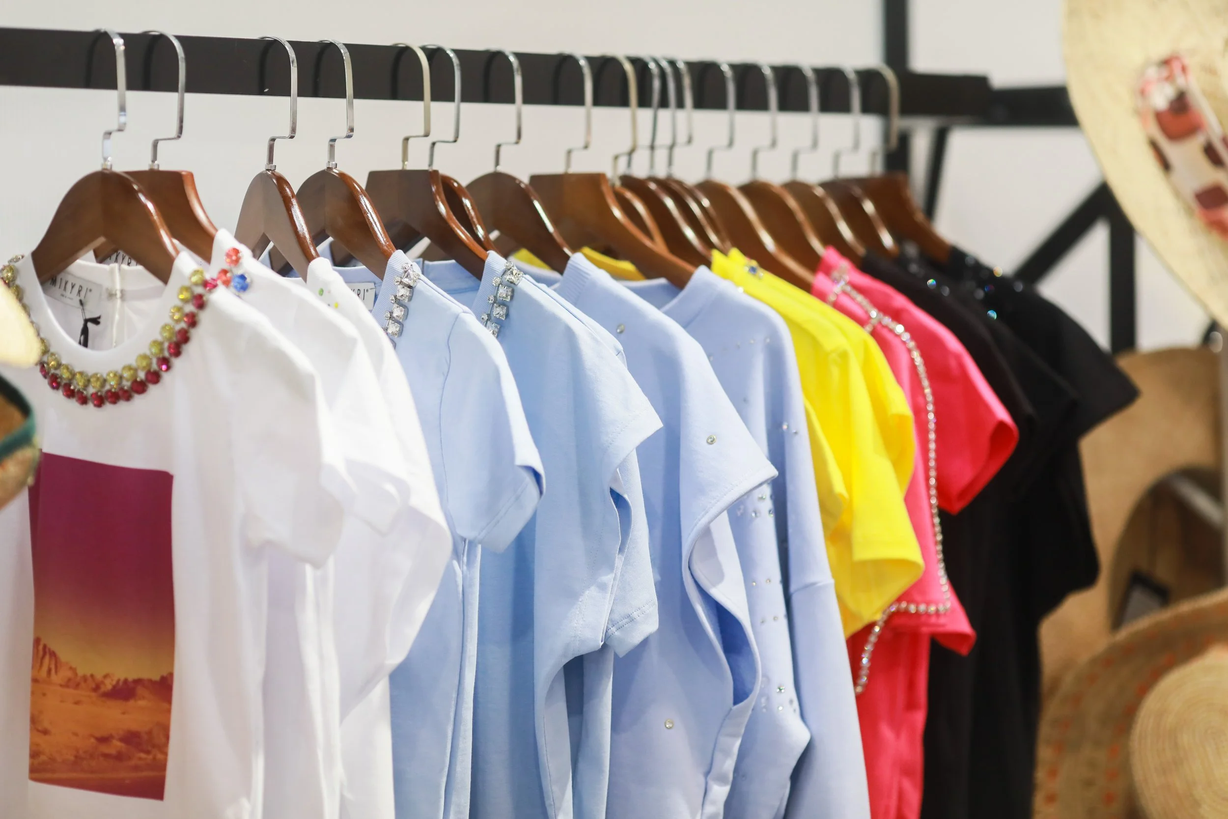A row of colorful T-shirts hanging on wooden hangers on a black clothing rack, including white, blue, yellow, pink, and black shirts, some decorated with beads or prints, in a store or boutique setting.