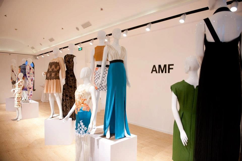 Display of mannequin dresses in an art gallery or showroom with spot lighting and a white wall featuring the letters 'AMF'.