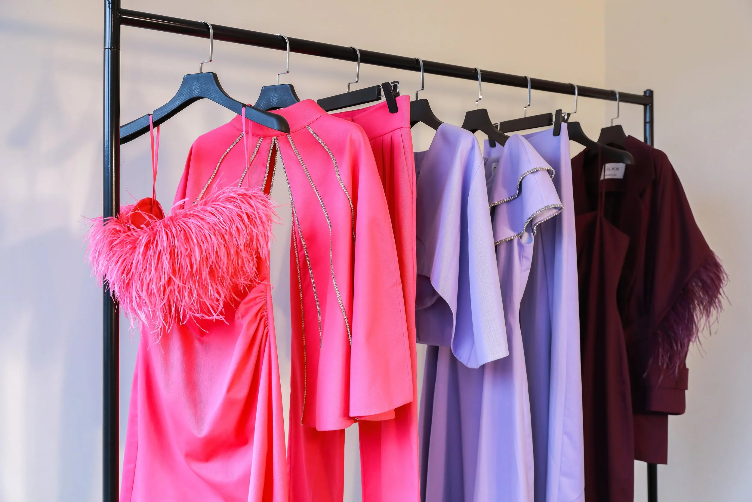 A clothing rack displaying colorful dresses in pink, lavender, and maroon, with the pink dress featuring feather details.