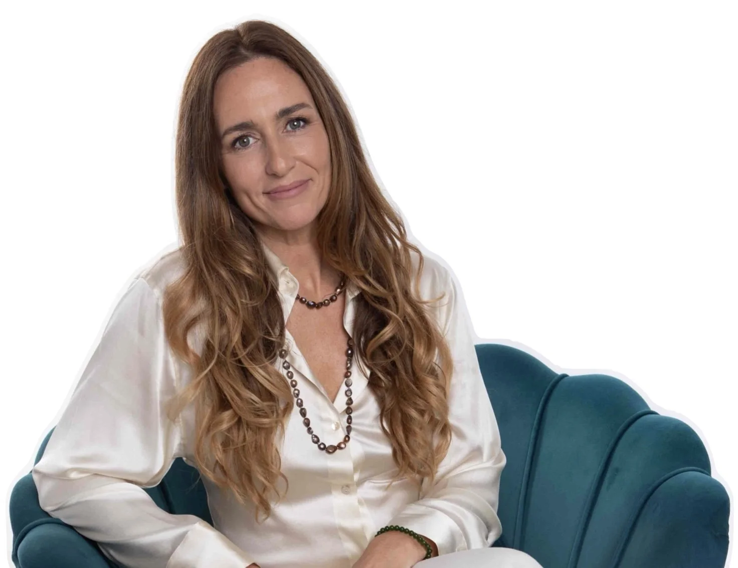 A woman with long, wavy auburn hair wearing a white silk shirt and a black beaded necklace, sitting on a teal velvet couch against a plain white background.