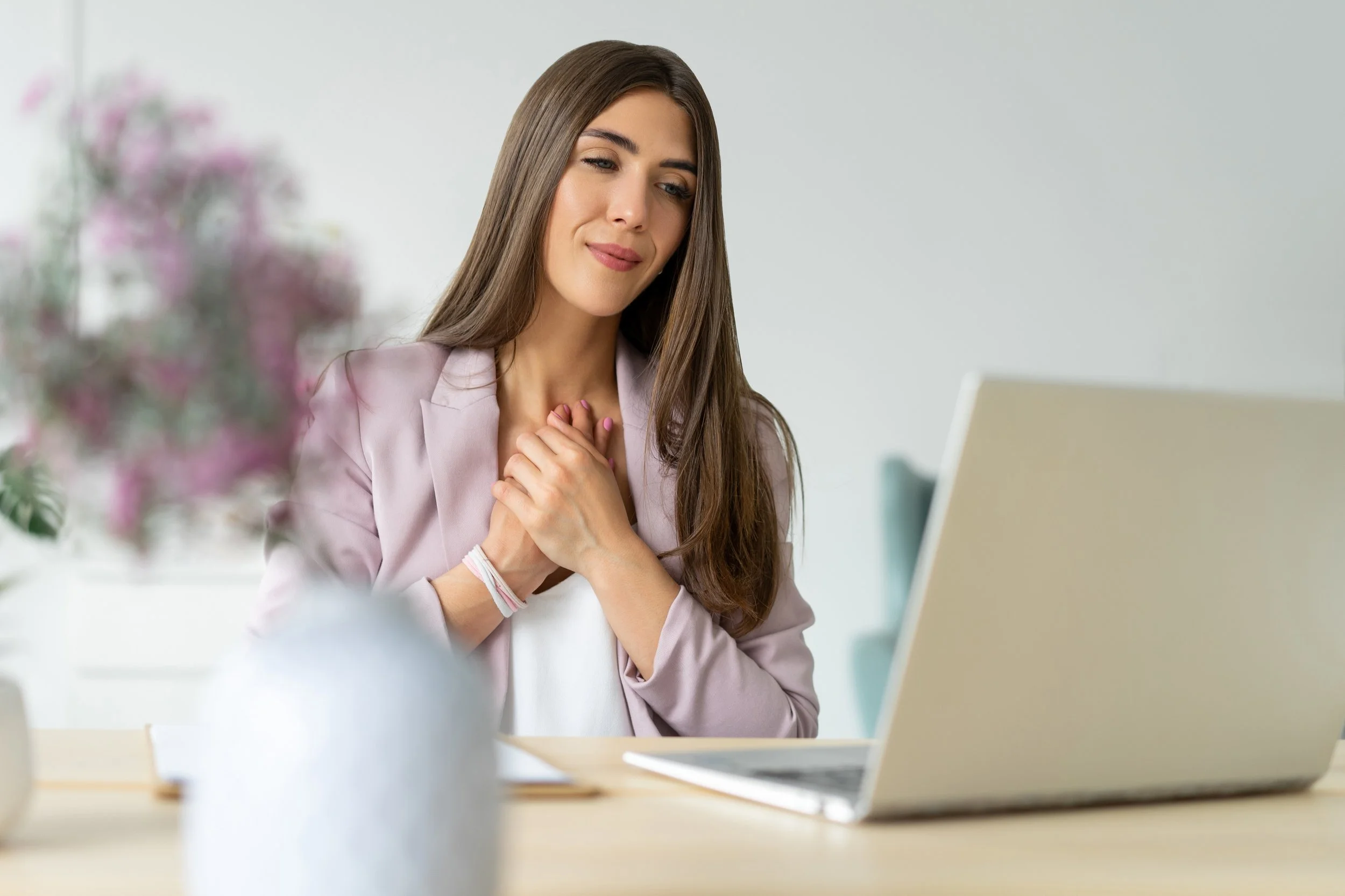 young woman with her hands on her heart dressed professionally looking at her laptop, representing. TSLT facilitator assisting her client with self love, empathy and compassionate connection