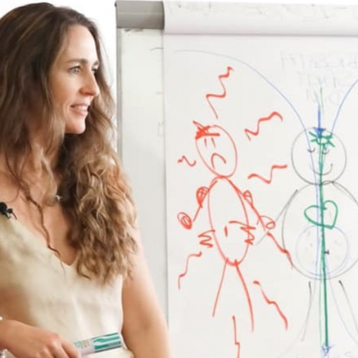 A woman with long, wavy hair standing next to a whiteboard with colorful drawings of a happy face, a heart, and various lines, smiling and looking to the side.