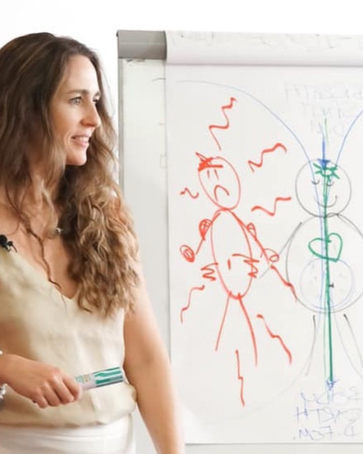 Tara Love Perry giving a presentation during a Transformational Self-Love Therapy™ training class, holding a marker pen, standing next to her diagram of a stressed person and a peaceful, self-embodied person.