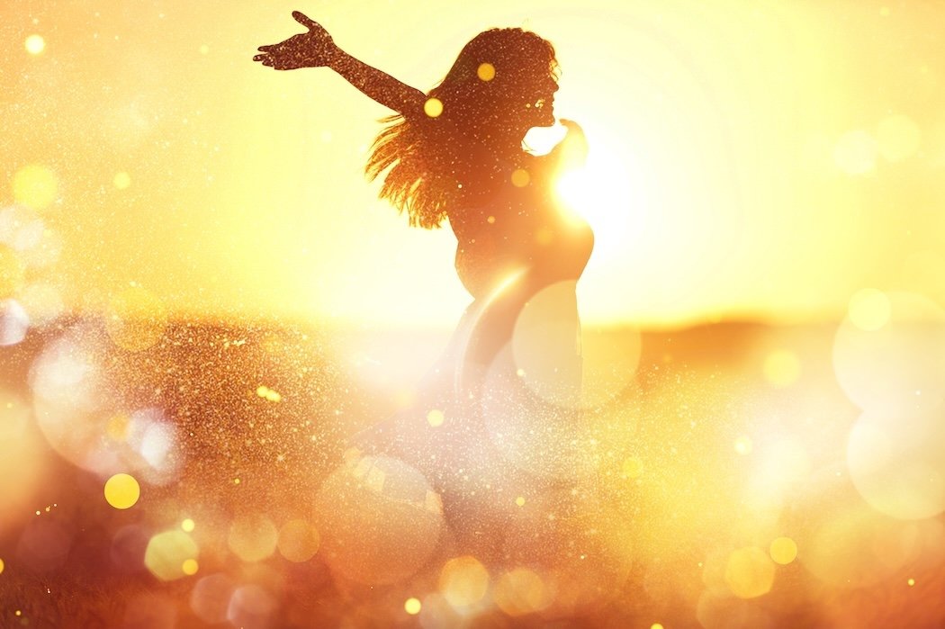A silhouette of a woman outdoors during sunset, with her arms raised, surrounded by a golden, glowing bokeh effect.