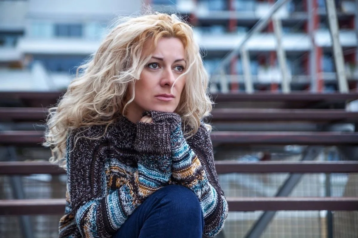A woman with curly blonde hair sitting on outdoor bleachers, resting her chin on her hand, looking thoughtful.