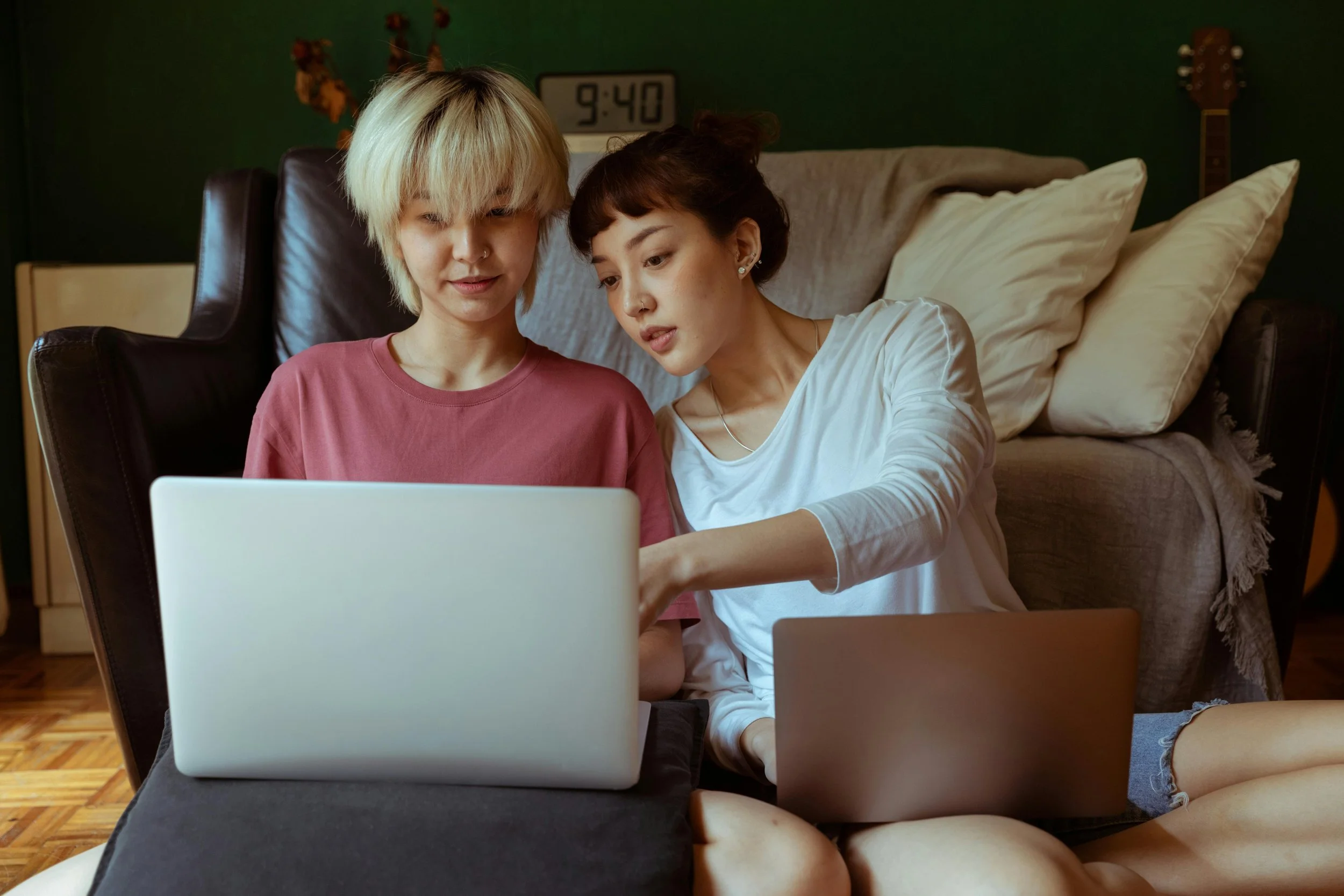 2 people sitting next to each other on the floor with laptops. One person is leaning over and pointing at the other persons laptop