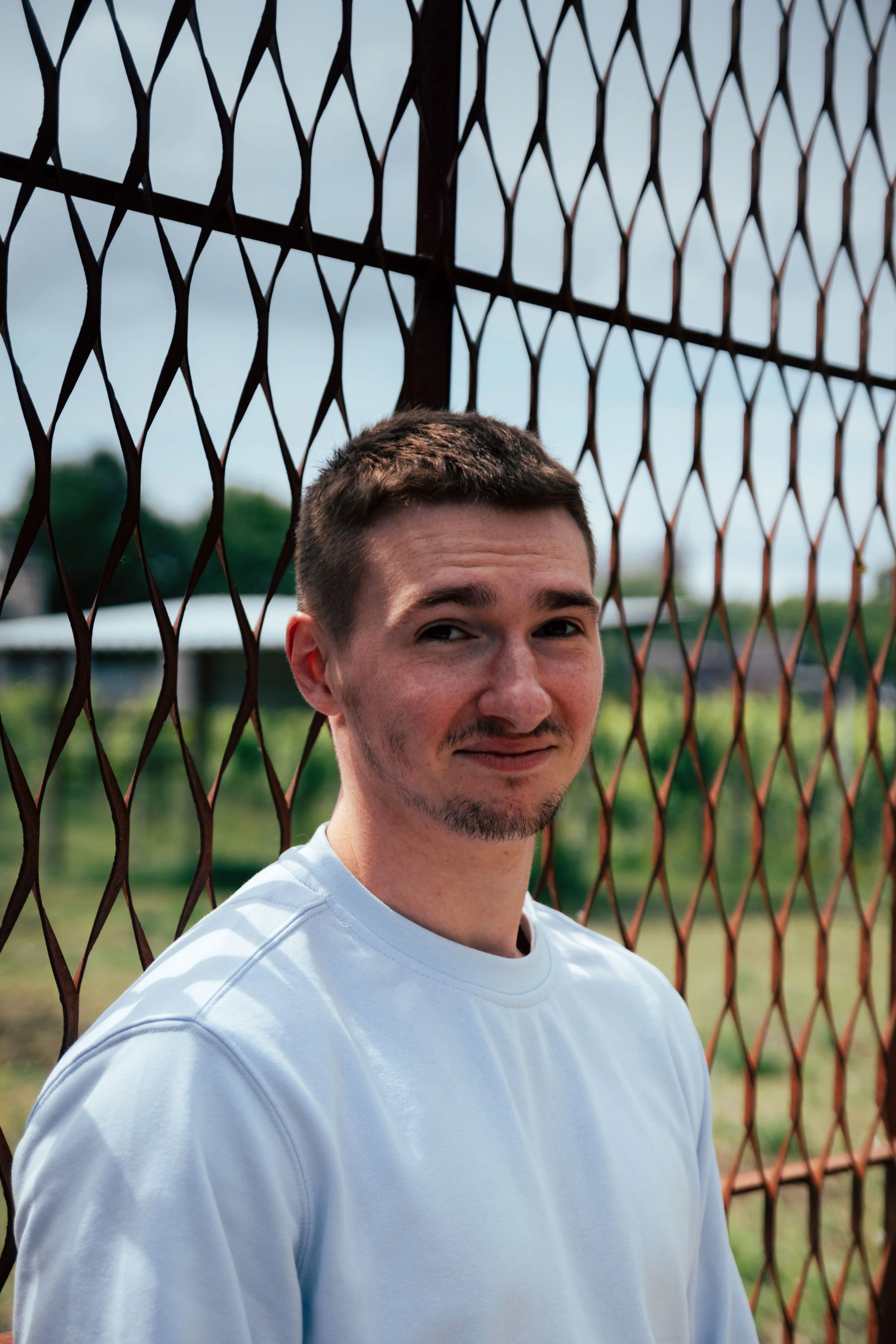 A man in a white t-shirt smiling, leaning against a fence