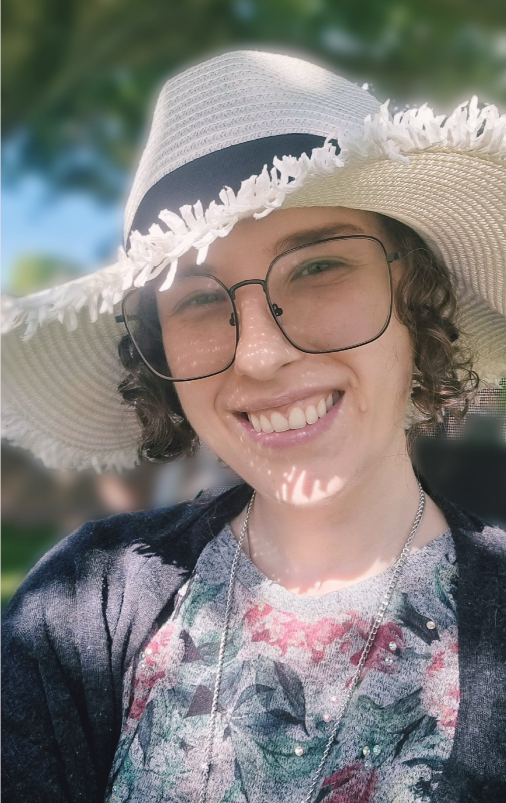 Max smiling outdoors wearing a white wide-brimmed straw hat, glasses, and a floral top.