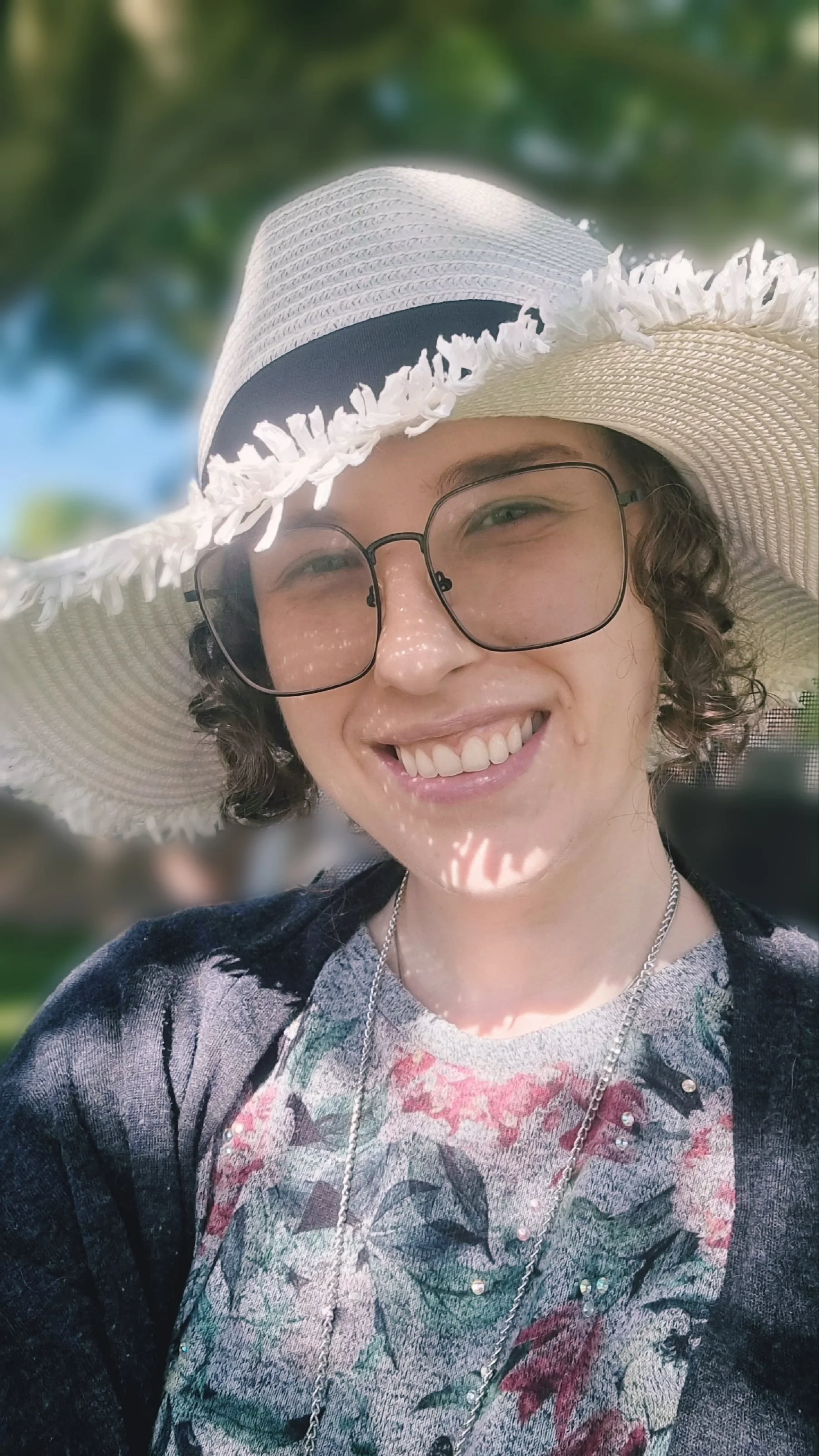 Max smiling outdoors wearing a white wide-brimmed straw hat, glasses, and a floral top.
