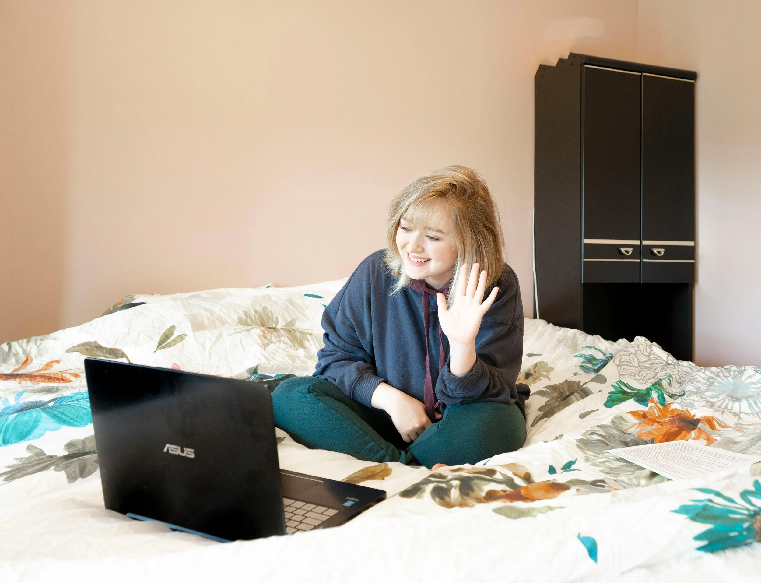 A young person waving at a laptop as they join a consultation from their bed.