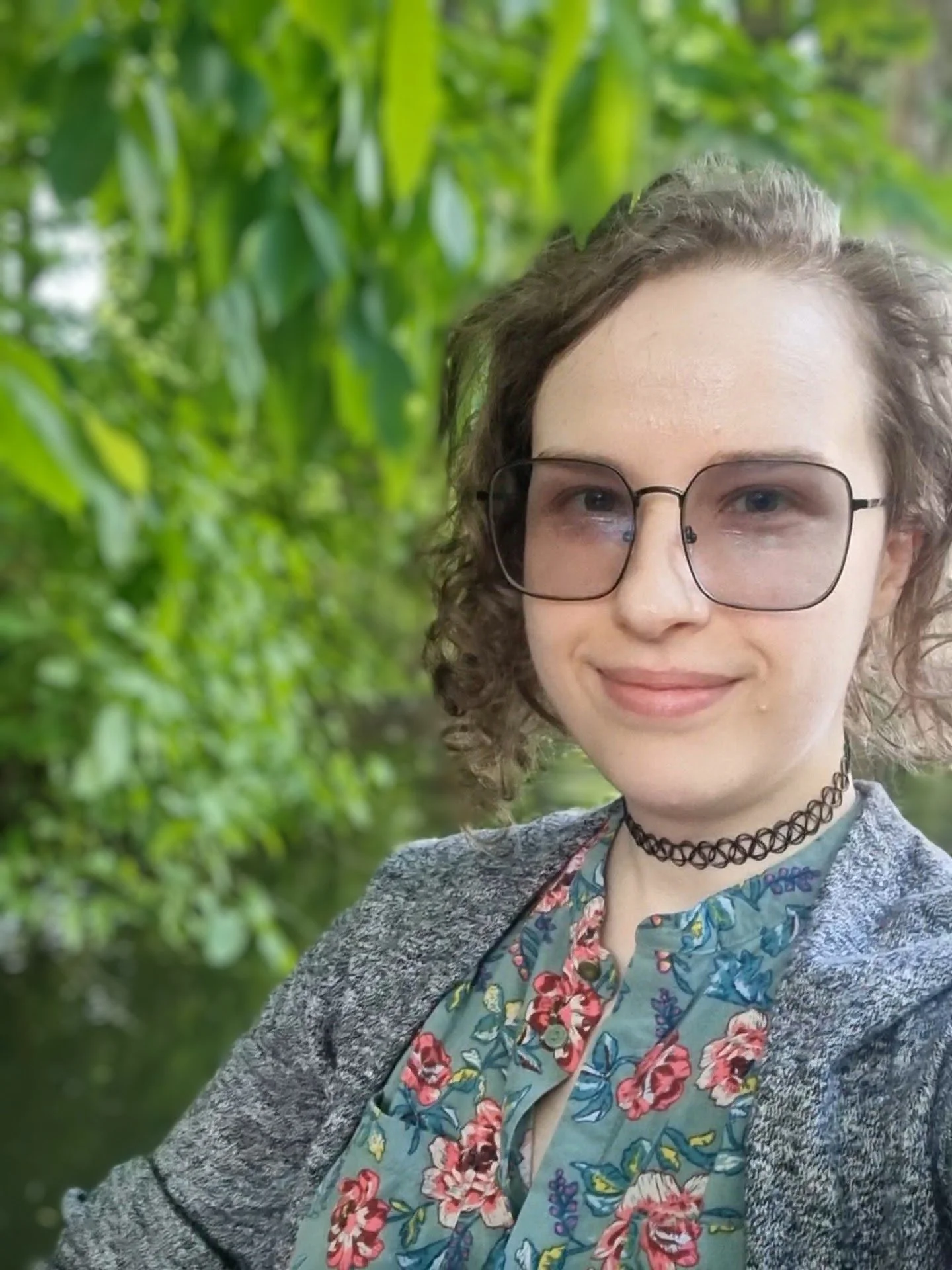Max standing outdoors in front of green foliage, smiling softly. They have curly brown hair and are wearing a floral shirt, a choker, a grey jacket, and glasses.