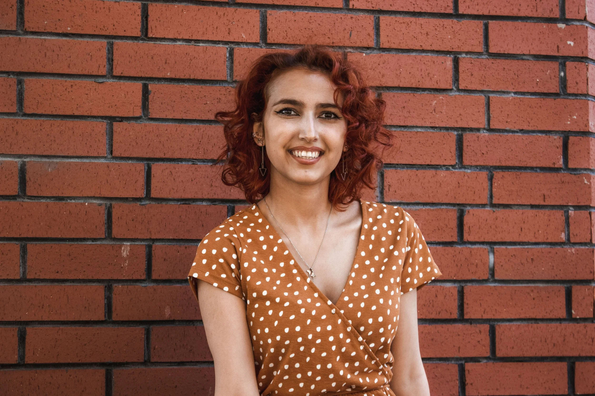 A woman with red curly hair against a brick wall, smiling