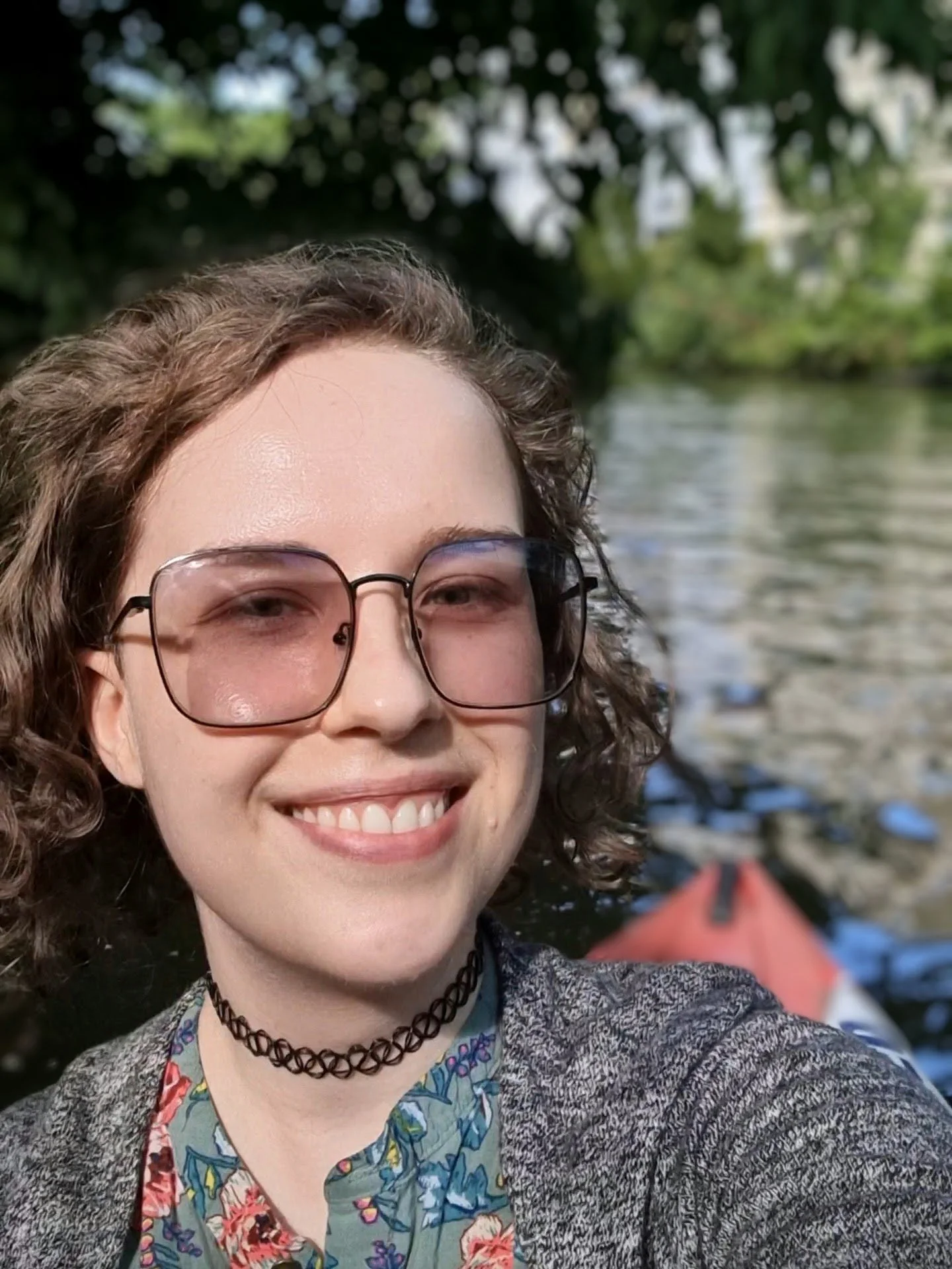 A person with curly brown hair wearing glasses, a floral shirt, and a choker necklace, smiling on a boat on a river with trees in the background.