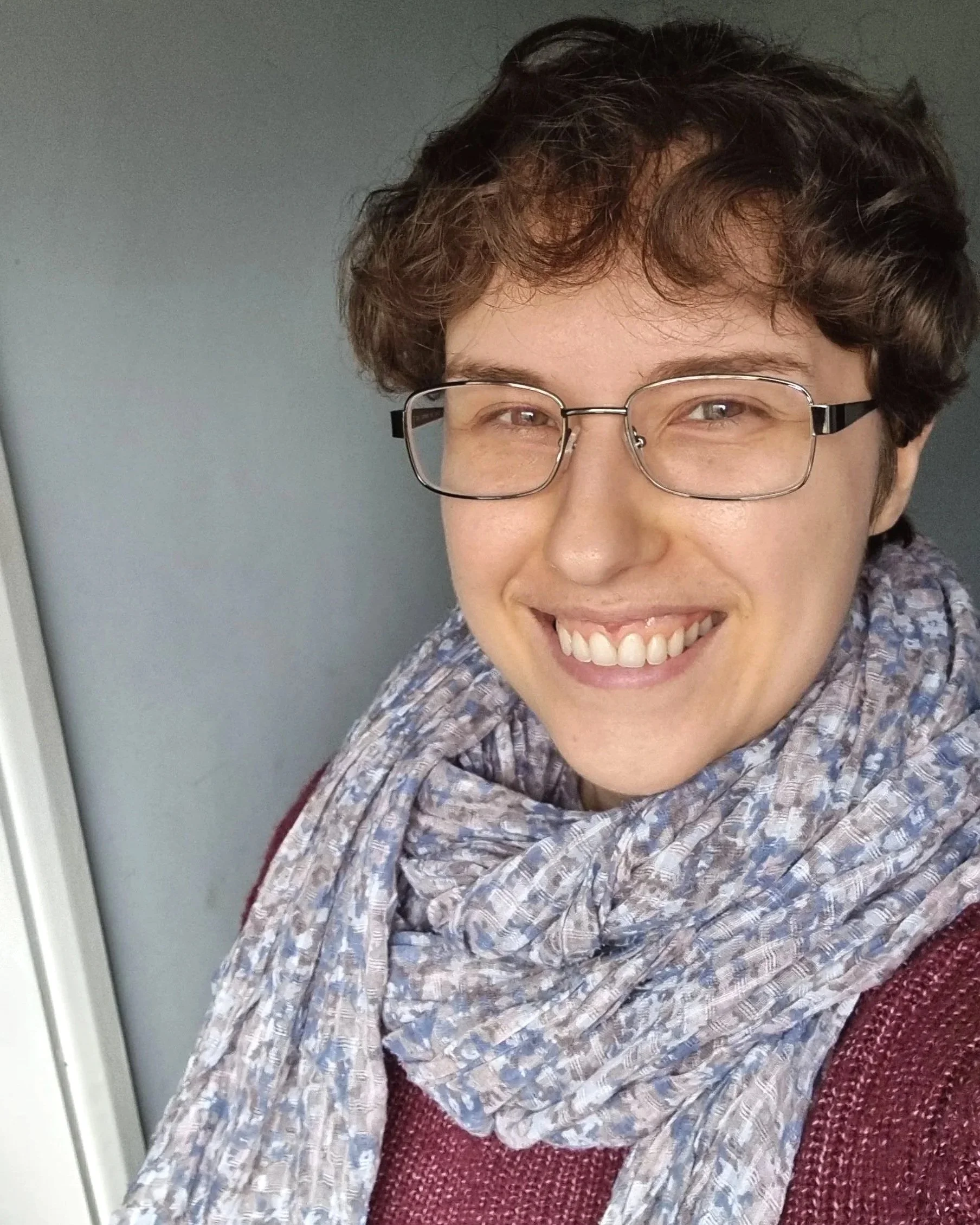 Max, smiling at the camera, wearing glasses, a grey and blue scarf, and a burgundy jumper.