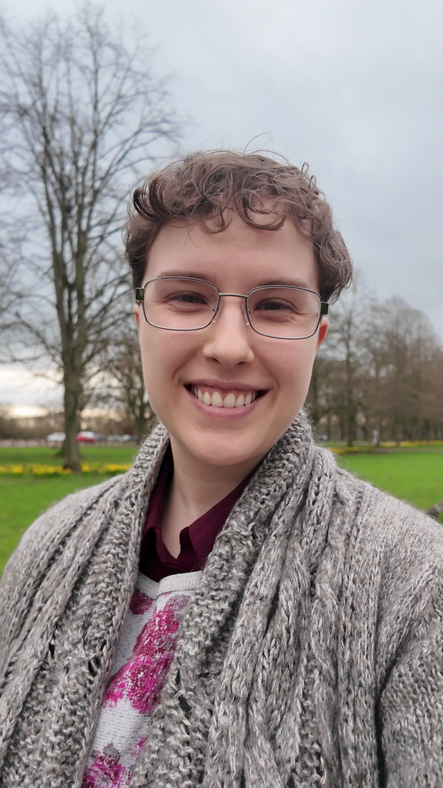 Max, smiling, with glasses and short curly hair, wearing a scarf and a maroon shirt, standing outdoors in a park with trees and green grass.