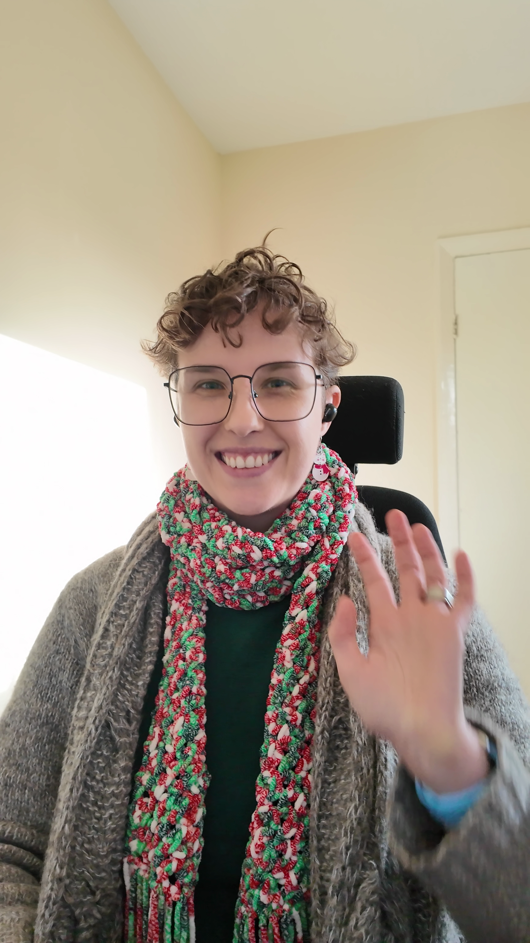 Max with short curly hair, glasses, and a big smile, wearing a patterned scarf, a gray cardigan, and waving at the camera.