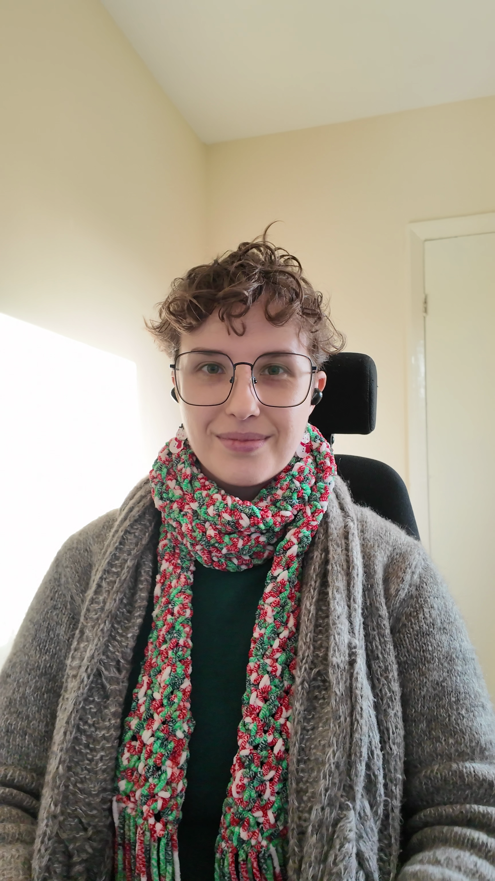 Max with short curly hair, wearing glasses, a colourful scarf, and a cosy sweater, sitting in a black chair in front of a beige wall and white door, smiling at camera