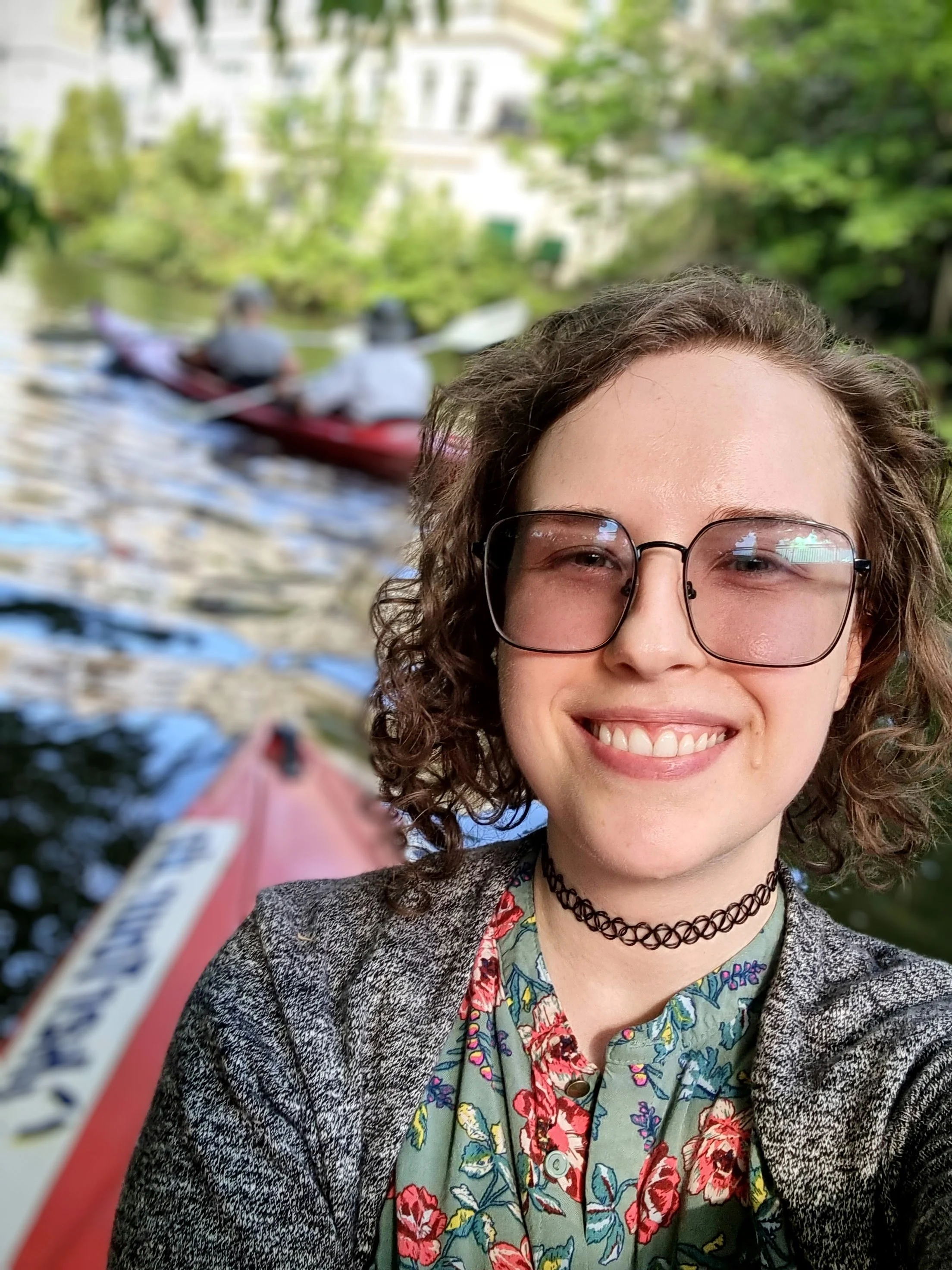 Max, with curly hair, wearing glasses, a choker, and a floral shirt, smiling for a selfie in front of a river with people kayaking and greenery in the background.
