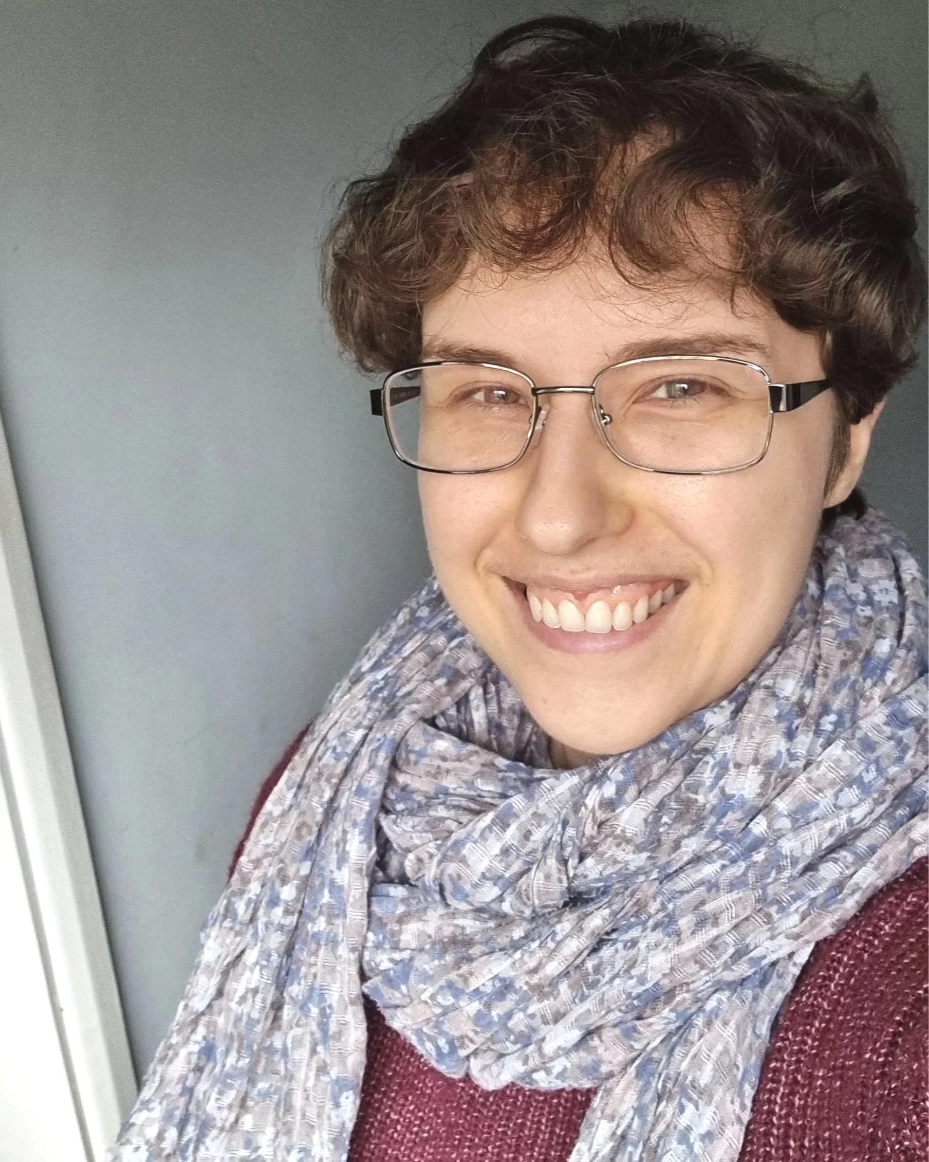 Close-up of Max, smiling, with short curly brown hair, wearing glasses, a patterned scarf, and a maroon sweater, against a grey background.