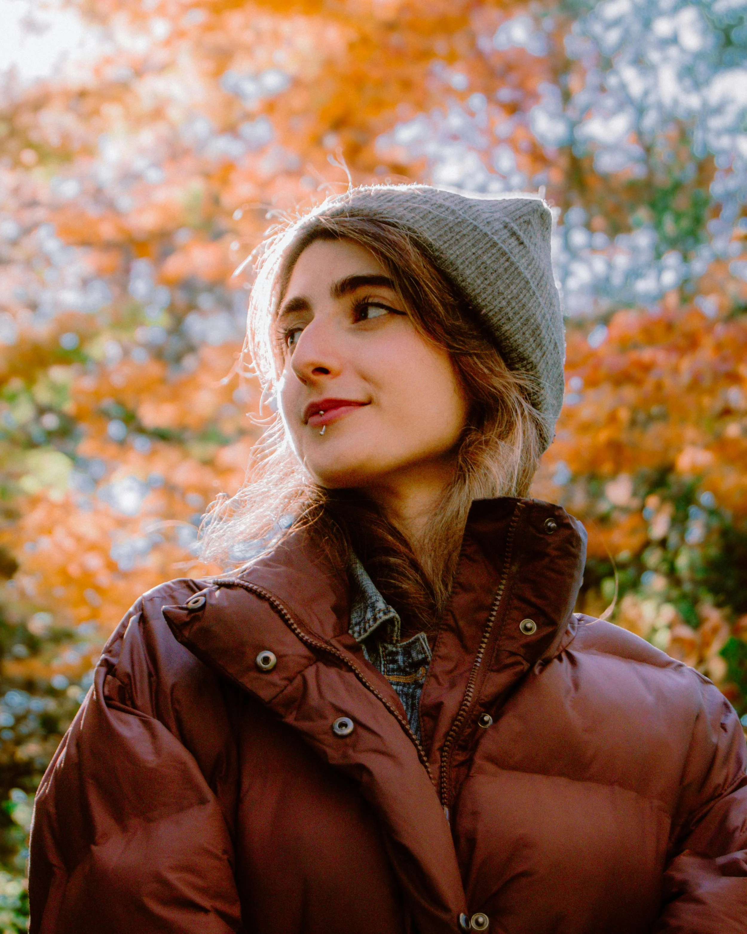 A happily smiling woman in a beanie, leather jacket, and lip piercing.