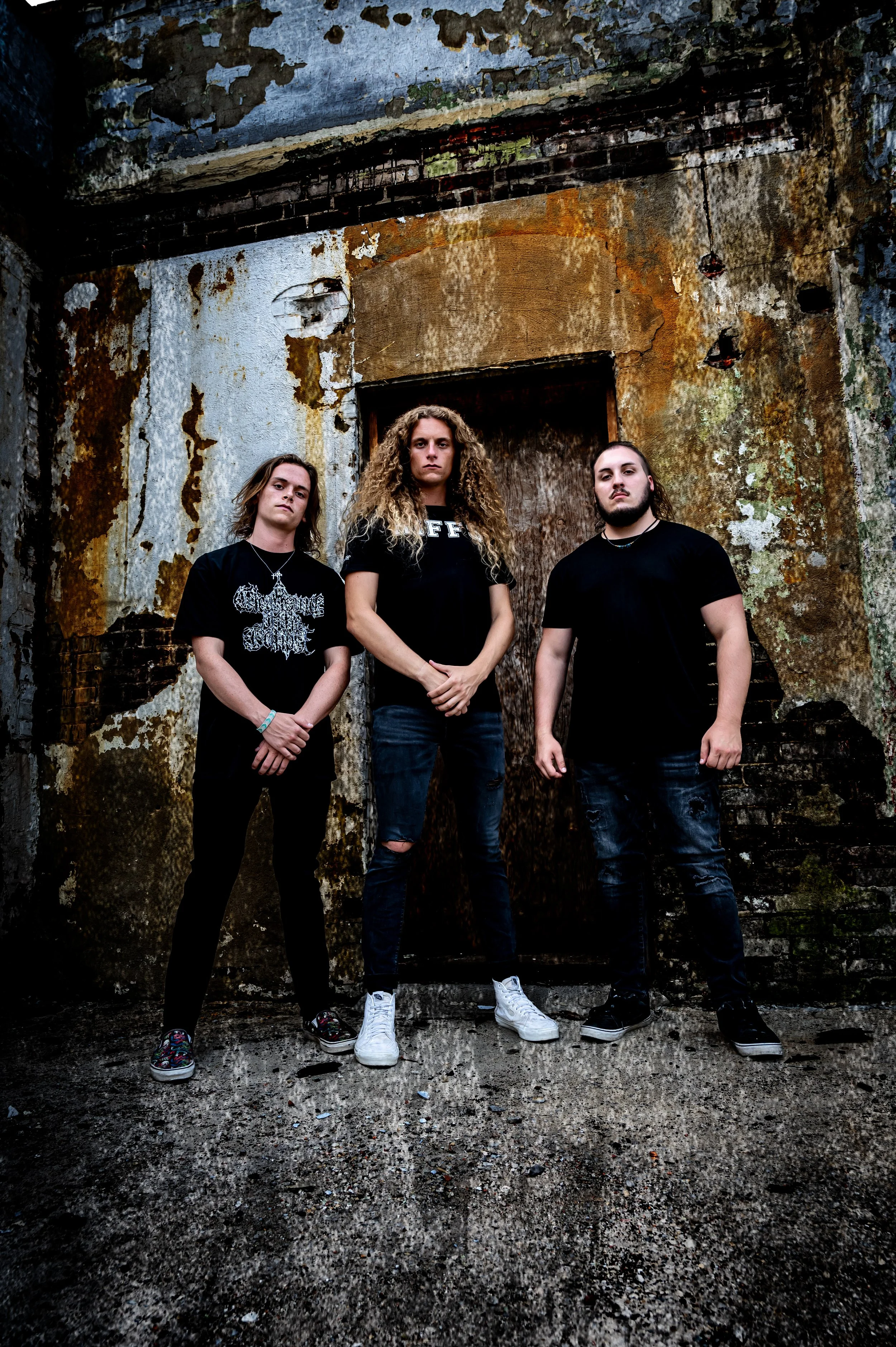 The members of Brotality standing in front of a dilapidated, rusted, and weathered wall with peeling paint and exposed brick, in an urban setting with a gritty atmosphere.