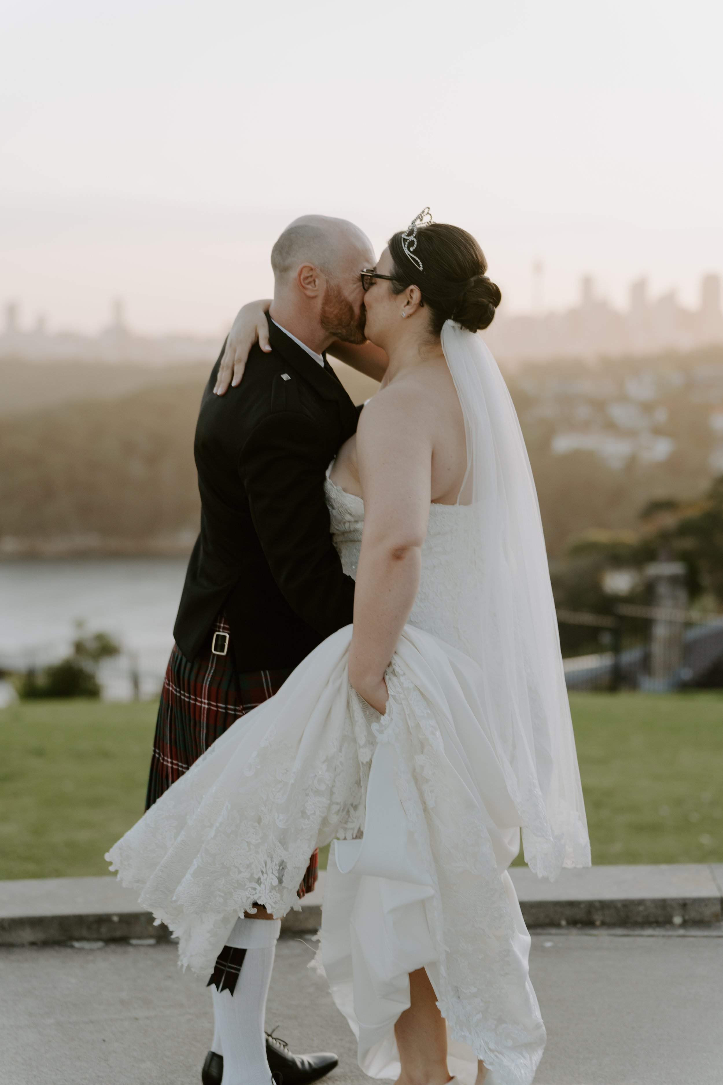 A bride and groom kissing outdoors during sunset, with city skyline in the background.