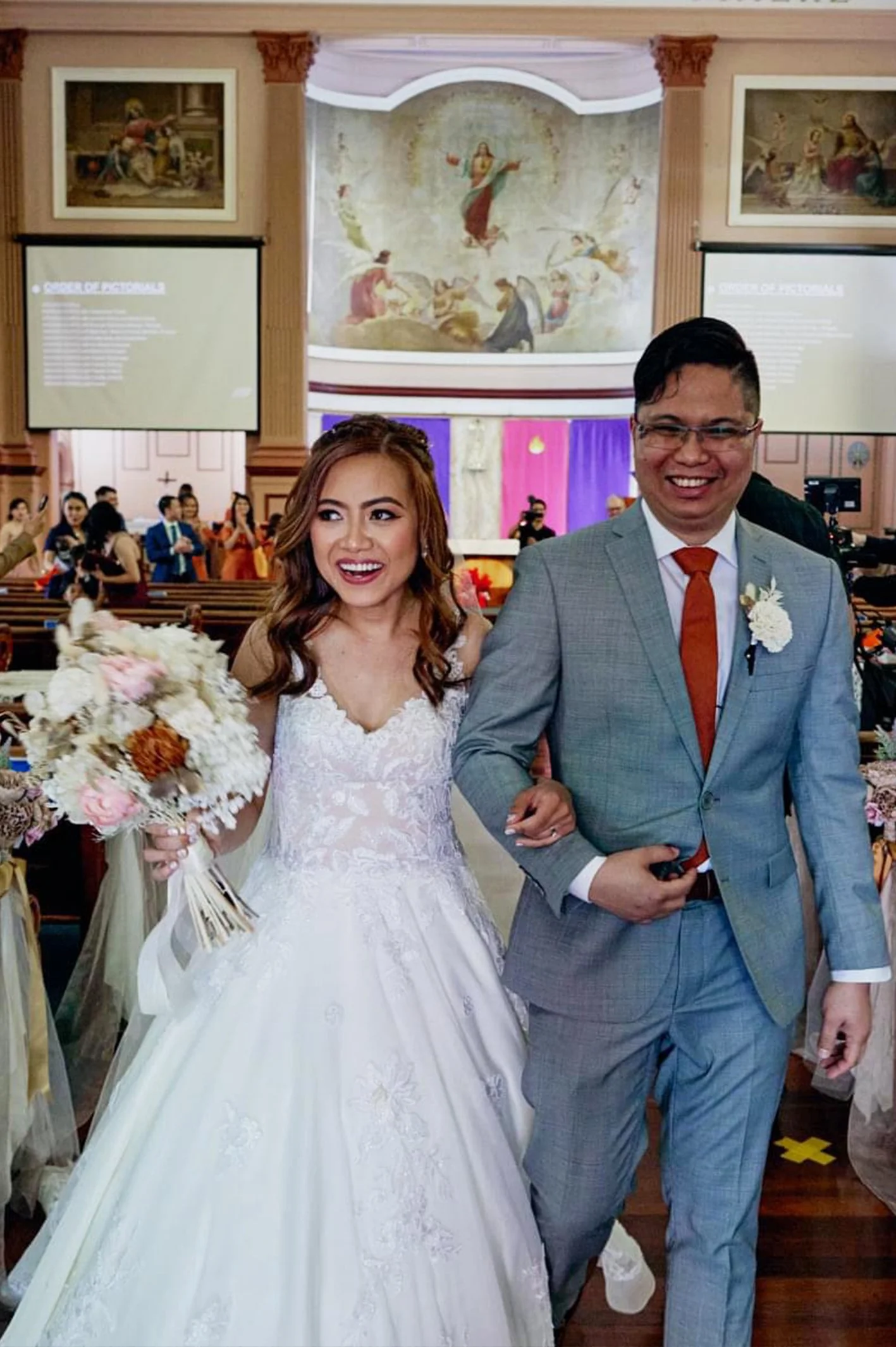 A bride and groom walking arm in arm inside a church during a wedding ceremony. The bride is holding a bouquet of flowers and is wearing a white wedding gown, and the groom is in a gray suit with a red tie.