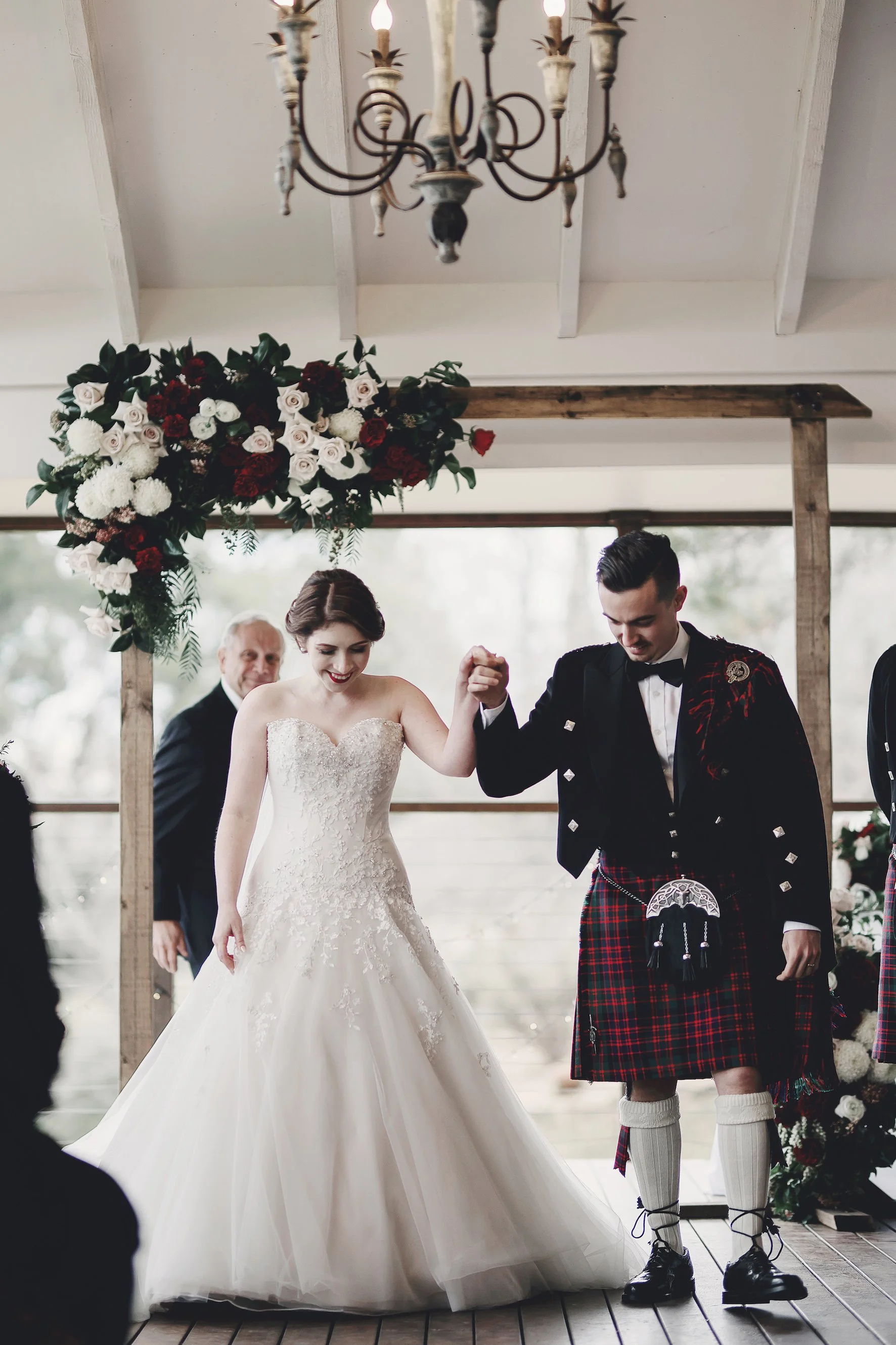 A bride and groom dancing at their wedding ceremony. The bride wears a strapless white wedding gown, and the groom wears a traditional Scottish kilt with a black jacket. They hold hands and smile as they dance, with a floral arrangement and an elderly man in the background.