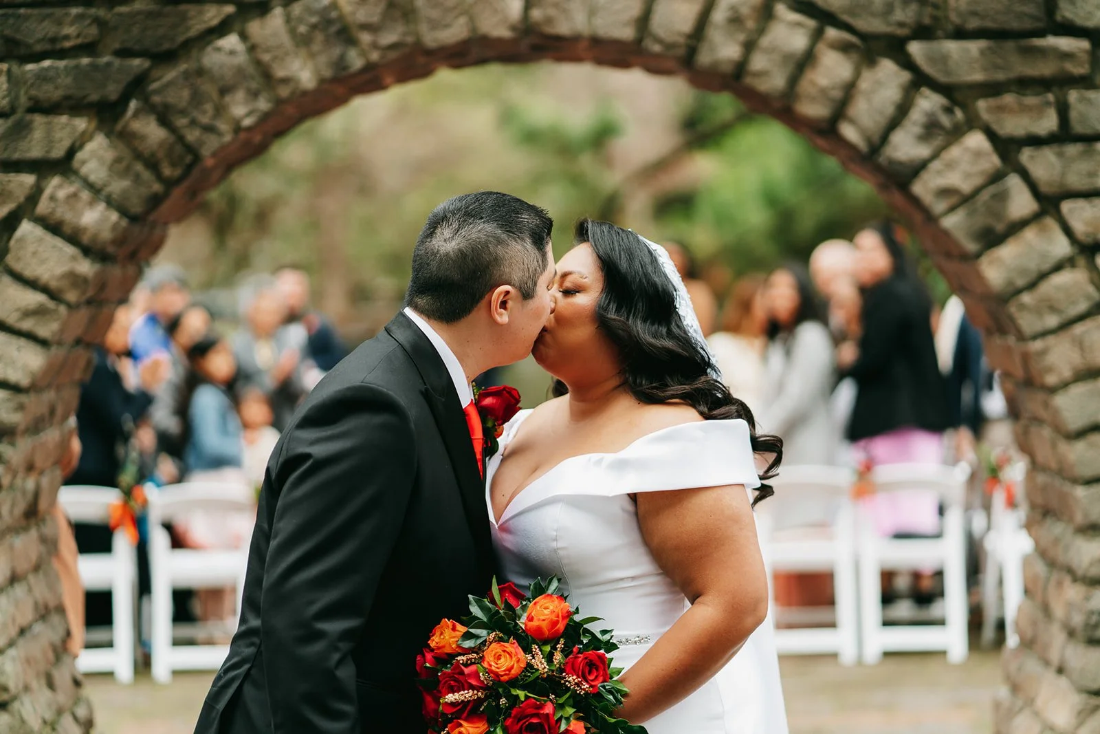 A bride and groom kiss under a stone arch during their wedding ceremony, with guests in the background.
