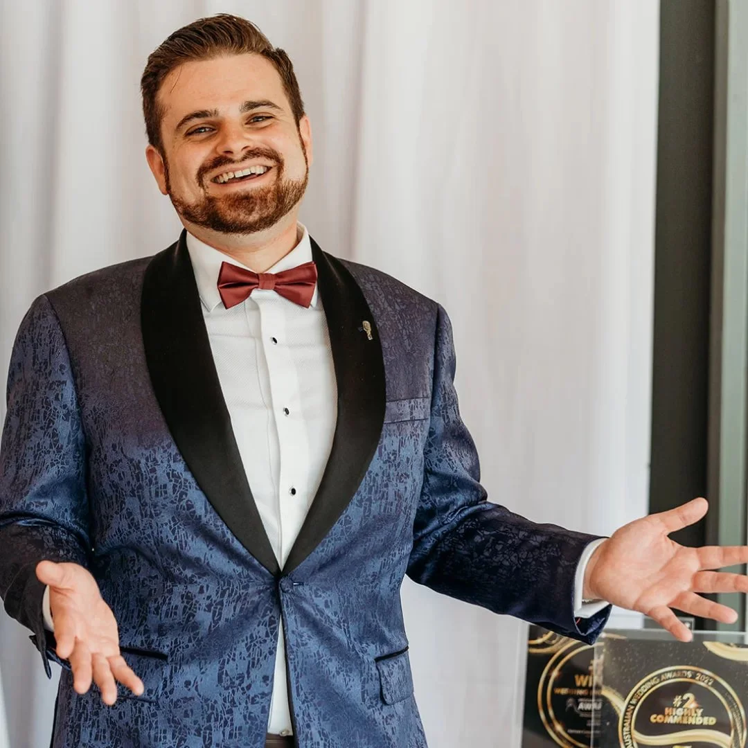 A man in a tuxedo with a red bowtie, smiling with arms outstretched, standing indoors in front of white curtains and a glass award.