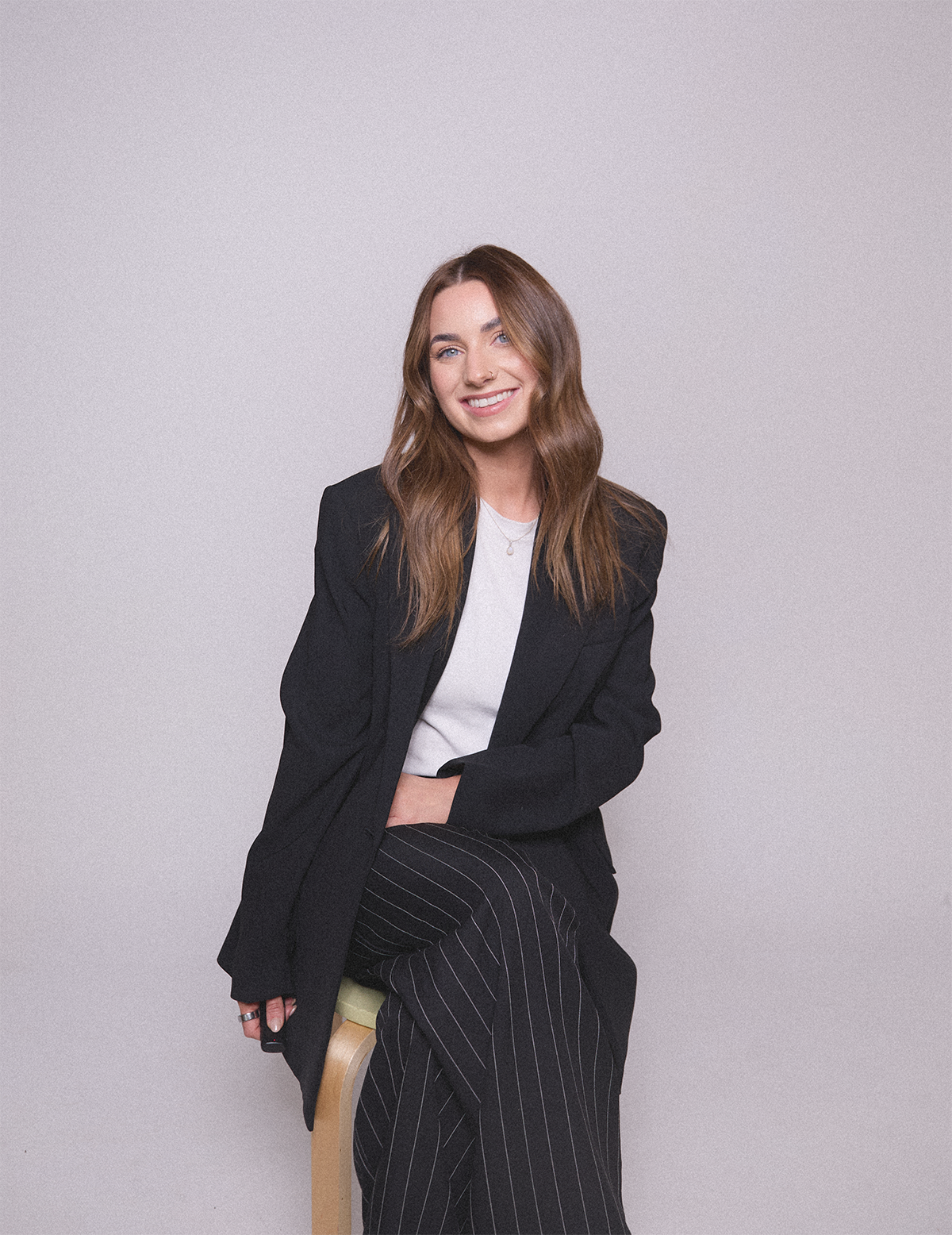 Emily Rose smiling and sitting on a stool against a plain light gray background, wearing a black blazer, white top, and black pinstripe pants.
