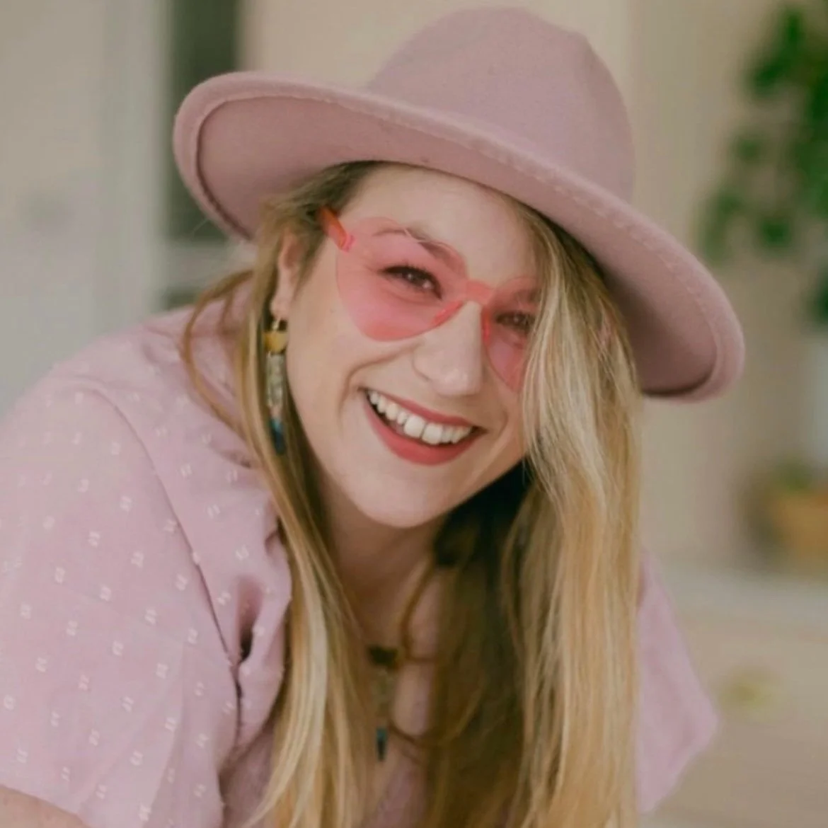 Woman smiling with pink sunglasses, pink wide-brimmed hat, and colorful earrings.